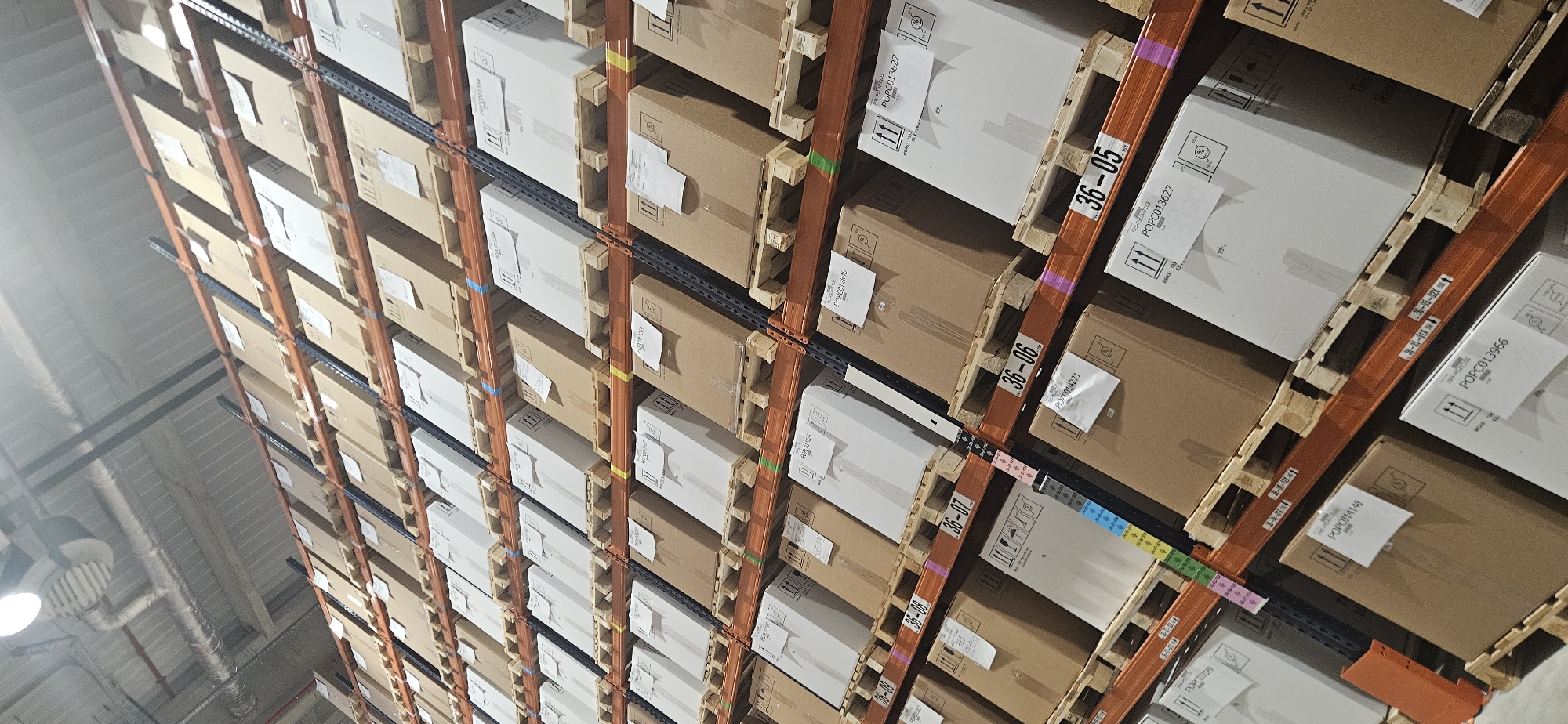 The image provides an upward-looking view within a large, organized warehouse or storage facility in Plovdiv, Bulgaria. The main subjects are rows of industrial metal shelving systems, densely packed with numerous cardboard boxes of varying sizes and colors (mostly brown and white) stacked on wooden pallets. No people are visible, focusing instead on the vast scale of the stored goods.
The scene depicts a static moment of inventory storage within a highly organized indoor setting. The overhead lighting fixtures and industrial ceiling structures are visible in the background, suggesting a brightly lit, active facility. The activity taking place is the efficient and systematic storage of packaged products, ready for distribution or retrieval.
Notable details include the multi-level racking system, which extends high upwards, indicating significant storage capacity. Colored bands (blue, green, yellow, pink) are visible on the vertical supports of the shelving, likely serving as visual cues for zone or aisle identification. Labels are affixed to both the boxes and the shelving beams.
Visible text from the labels includes specific location identifiers on the racks such as "36-09", "36-08", "36-07", "36-06", and "36-05". Product or inventory codes are prominently displayed on box labels, including "POPC012840", "POPC013627", "POPC014271", "POPC014148", and "POPC013966". Other text snippets like "MEAS: 115", "705-PSLGT103", "11" (likely a "this way up" symbol), "This", and "Thay" are also discernible. The overall impression is one of a meticulously managed and extensive logistics operation.