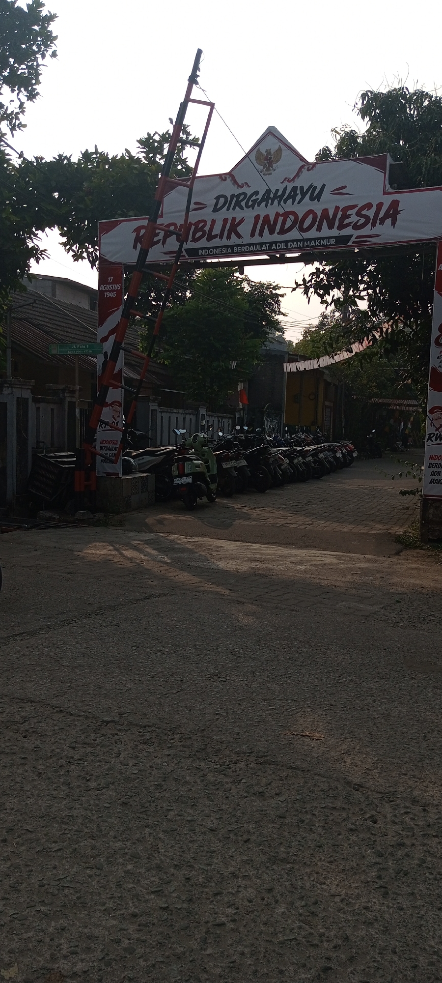 The image depicts an outdoor scene in what appears to be a residential or communal area in Setu, Indonesia, likely near a gate or entrance. Dominating the upper part of the frame is a banner that reads "DIRGAHAYU REPUBLIK INDONESIA" with the Garuda Pancasila emblem at its apex, celebrating Indonesian Independence. Below this, smaller text states "INDONESIA BERDAULAT ADIL DAN MAKMUR." To the left of the banner, a striped barrier, typical of those found at entrances or checkpoints, is partially lowered.
The ground is paved with asphalt and shows tire marks and debris. A row of parked motorcycles is visible, suggesting a parking area. Some of the motorcycles are scooters, and one has a license plate that appears to read "B 6252 YFF." To the left of the motorcycles and barrier, a green street sign is mounted on a pole, indicating "Jl. Pata 1" with what looks like a postal code or street identifier. Behind the barrier and motorcycles, there are buildings with tiled roofs and fences, characteristic of Indonesian architecture. Lush green trees and foliage surround the area, and the sky is a bright, overcast white, suggesting it is daytime. The overall impression is a functional, everyday setting decorated for a national celebration.