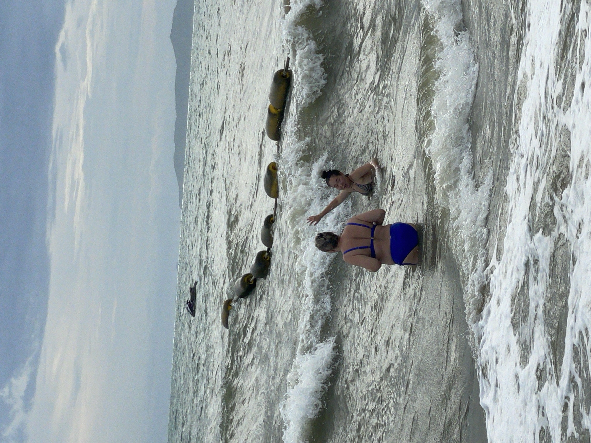 The image depicts two individuals in the ocean at Pattaya City, Thailand. The person in the foreground, viewed from behind, is wearing a blue bikini and is standing in waist-deep water. To their right, another person is in the water, appearing to be swimming or playing, wearing a patterned bikini. Their left arm is raised. A line of yellow buoys stretches across the water in the middle ground. In the distance, a jet ski is visible on the water, and landmasses can be seen on the horizon. The sky is overcast with clouds. The water is choppy, with waves breaking on the shore in the foreground.