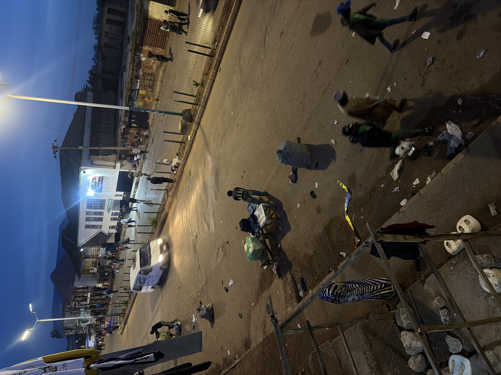In Bukuru, Nigeria, a street scene unfolds at dusk. A white van is in motion on the asphalt road, its headlights illuminated. Several people are visible walking along the sidewalk and crossing the street. Some are carrying items, and others appear to be engaged in conversation. The street is lined with buildings, some of which have shops or market stalls. Various objects are scattered on the ground, including trash and some goods displayed on a table. The sky is a deep blue, indicating the transition from daylight to night, with streetlights beginning to cast their glow. The overall impression is of an active street in a Nigerian city as evening approaches.