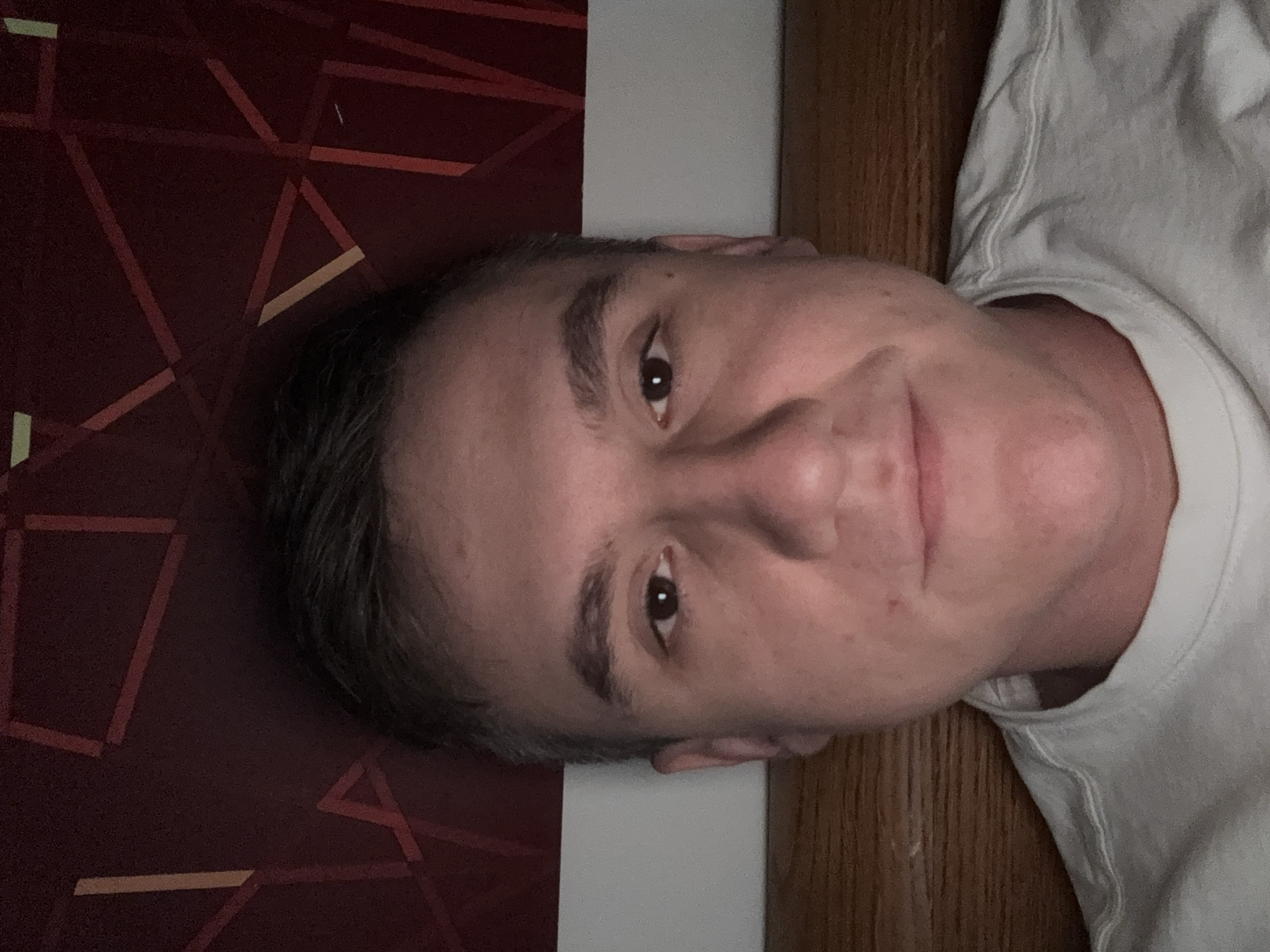 A man with short, dark hair is facing the camera. He has brown eyes and is wearing a white t-shirt. He is positioned against a backdrop of a wall with a geometric pattern in reddish-brown and a section of wood paneling. The location is not provided.