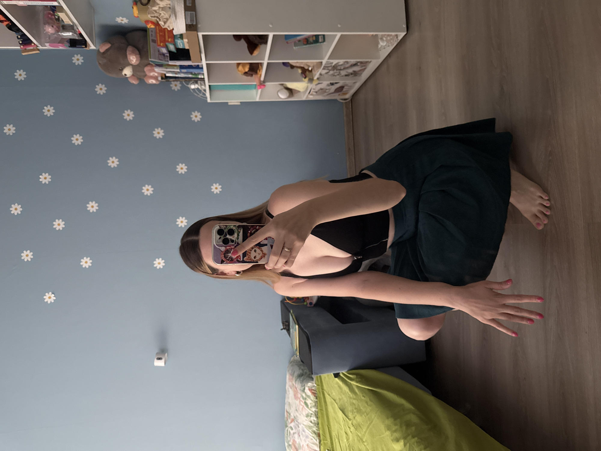 The image shows a young woman crouching on a wooden floor in what appears to be a bedroom. She is taking a selfie with her phone, which has a colorful case featuring cats. She is wearing a black, zip-up crop top and a dark green skirt. Her fingernails are painted pink. In the background, there is a bed with green and floral bedding, a blue wall decorated with white daisy stickers, a shelf unit filled with various items including stuffed animals and books, and a portion of a wardrobe. The lighting suggests an indoor setting during the daytime. The overall impression is a casual, personal space.