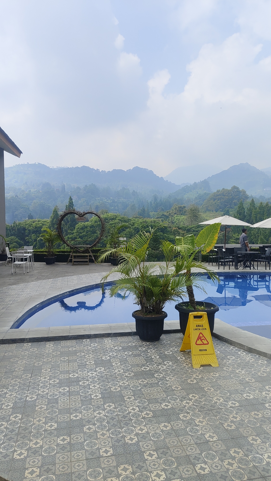 The image depicts a swimming pool area with a scenic mountain backdrop, likely located in Cisarua, Indonesia. In the foreground, a blue swimming pool is visible, with its reflection showing a heart shape. The pool deck is paved with grey and white patterned tiles. Two potted plants, one a palm and the other a larger leafy plant, are placed beside the pool. A yellow caution sign warning of a slippery floor is also present. In the mid-ground, a large heart-shaped decorative structure made of wood and branches stands against a backdrop of lush green trees and rolling mountains shrouded in mist. A few tables and chairs are set up near the pool, with umbrellas providing shade. A person in a grey uniform is walking in the background near the tables. The sky is overcast with scattered clouds.