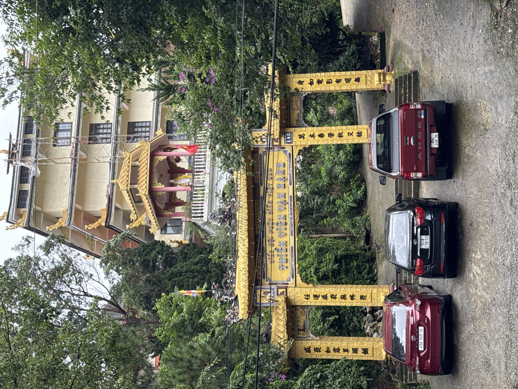 An outdoor scene in Lâm Viên - Đà Lạt, Vietnam, captures the entrance to a temple or monastery, likely Thien Vien Van Hanh Pagoda. A prominent golden archway, adorned with intricate designs and calligraphy in both Vietnamese and Chinese, serves as the main gateway. The Vietnamese text "THIÊN VIÊN VẠN HẠNH" is clearly visible on the archway's horizontal beam. Other Chinese inscriptions translate roughly to philosophical Buddhist verses about impermanence, such as "Body like a movie, existent yet non-existent" and "Creepers flourish in spring, wither in autumn."
Three cars are parked on the concrete path in front of the archway: a red Ford Ranger pickup truck (license plate 49C 248.38), a black sedan (license plate 51A 238.50), and a dark red SUV/crossover (license plate 49A-345.16). The "49" prefix on the plates confirms their origin from Lâm Đồng province, where Đà Lạt is located. Steps lead upwards behind the vehicles and through the archway.
Beyond the archway, a multi-story building with a light-colored facade and multiple windows is visible. Part of a traditional golden temple roof with ornate details can be seen near this building. Scaffolding is present on the left side of the building, indicating ongoing construction or maintenance. A small red flag and colorful Buddhist prayer flags are also visible among the trees.
The scene is lush with green trees and foliage, some featuring purple flowering plants, suggesting a verdant, natural setting. Overhead power lines cross the frame. The lighting indicates daytime, possibly overcast, creating a serene and tranquil atmosphere. No people are visible, but the parked cars suggest visitors or residents are present at the temple.