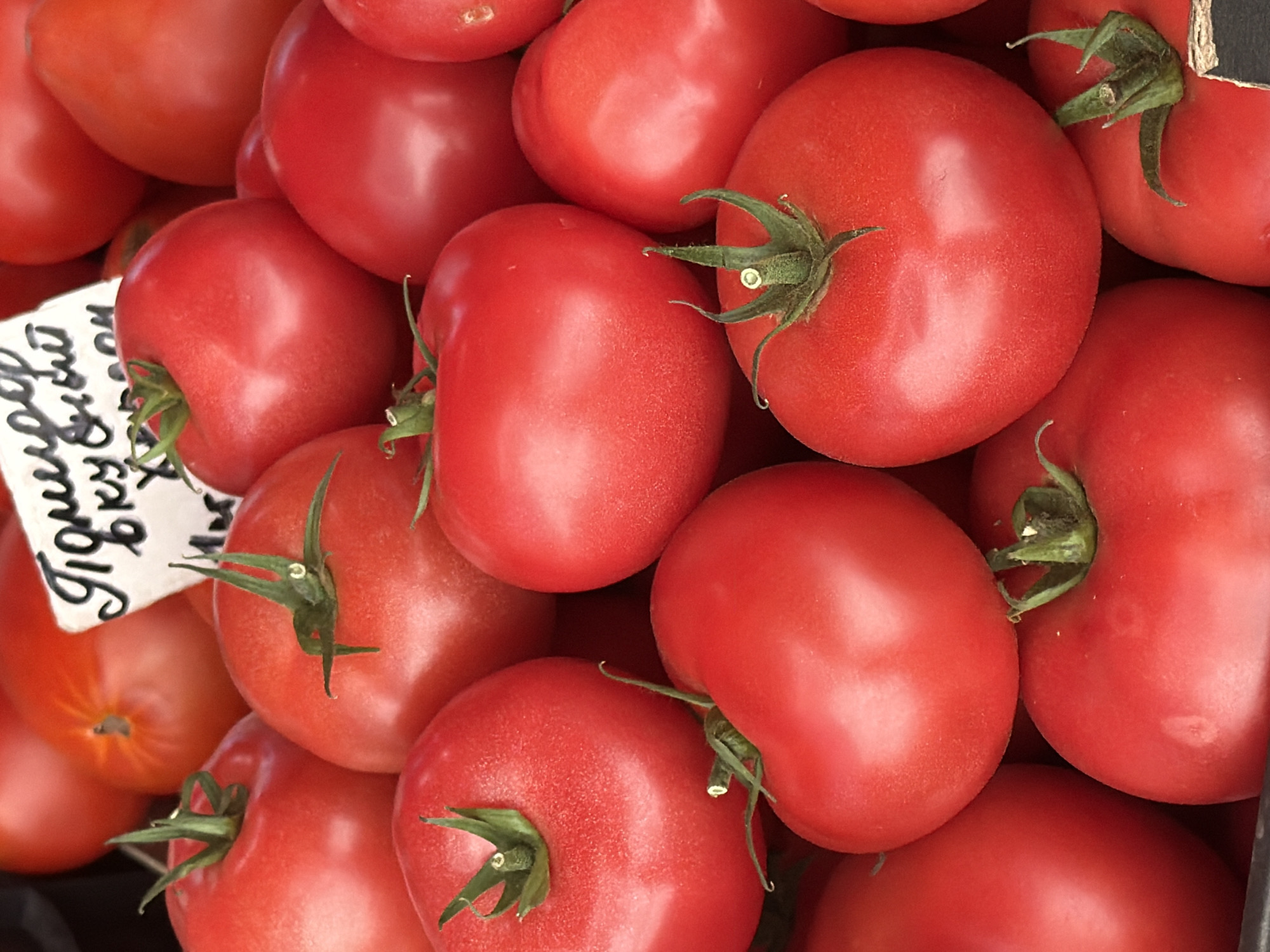 This media file features a close-up, overhead shot of a pile of ripe, red tomatoes. The tomatoes are the primary subjects, filling the frame. They appear to be fresh and ready for sale, likely at a market. The lighting suggests it could be daytime, possibly indoors or in an area with diffused sunlight, as there are no harsh shadows.
There is a white tag with handwritten text in Cyrillic script visible in the upper left quadrant of the image. The text appears to be a price or description related to the tomatoes, possibly from a market stall in Minsk, Belarus, as indicated by the location context. The tomatoes themselves have green stems and sepals, indicating they are recently harvested. The overall impression is one of abundance and freshness. No people are visible in the frame, and the focus is solely on the produce. The setting appears to be a market or grocery display.