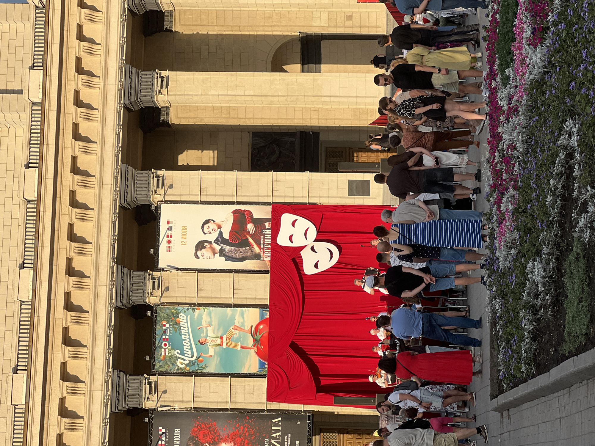 The image depicts an outdoor event in front of the Novosibirsk Opera and Ballet Theatre in Novosibirsk, Russia. A temporary stage with a red curtain backdrop has been set up. On the stage, a small ensemble of musicians is performing, with individuals playing string instruments and an accordion. A crowd of people are gathered in front of the stage, watching the performance. Large posters advertising upcoming performances are displayed on the facade of the theatre. The architecture of the theatre is visible, characterized by its classical style with columns and ornate detailing. The scene is illuminated by sunlight, casting shadows on the building and the ground. A flowerbed with various plants is in the foreground.