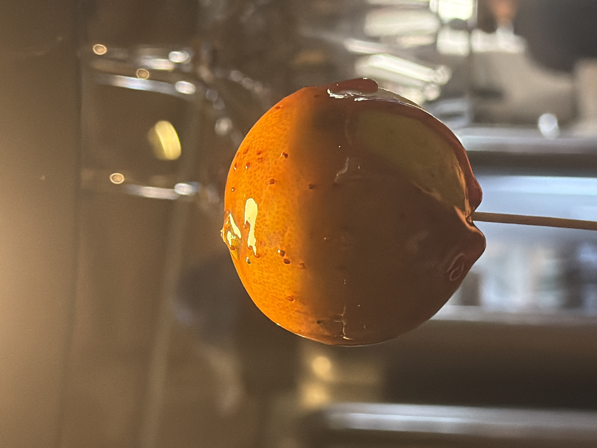 A close-up, eye-level shot shows a bright orange citrus fruit on a wooden skewer, partially coated in a glossy, dark brown caramel or chocolate sauce. The fruit's skin is textured with small, dark pores, and the sauce drips downwards, creating a wet, glistening effect. The background is softly blurred, featuring indistinct shapes of glassware and warm lighting, suggesting an indoor setting such as a restaurant or bar. There are no people or specific landmarks visible to indicate the city of Palmanova, Spain.