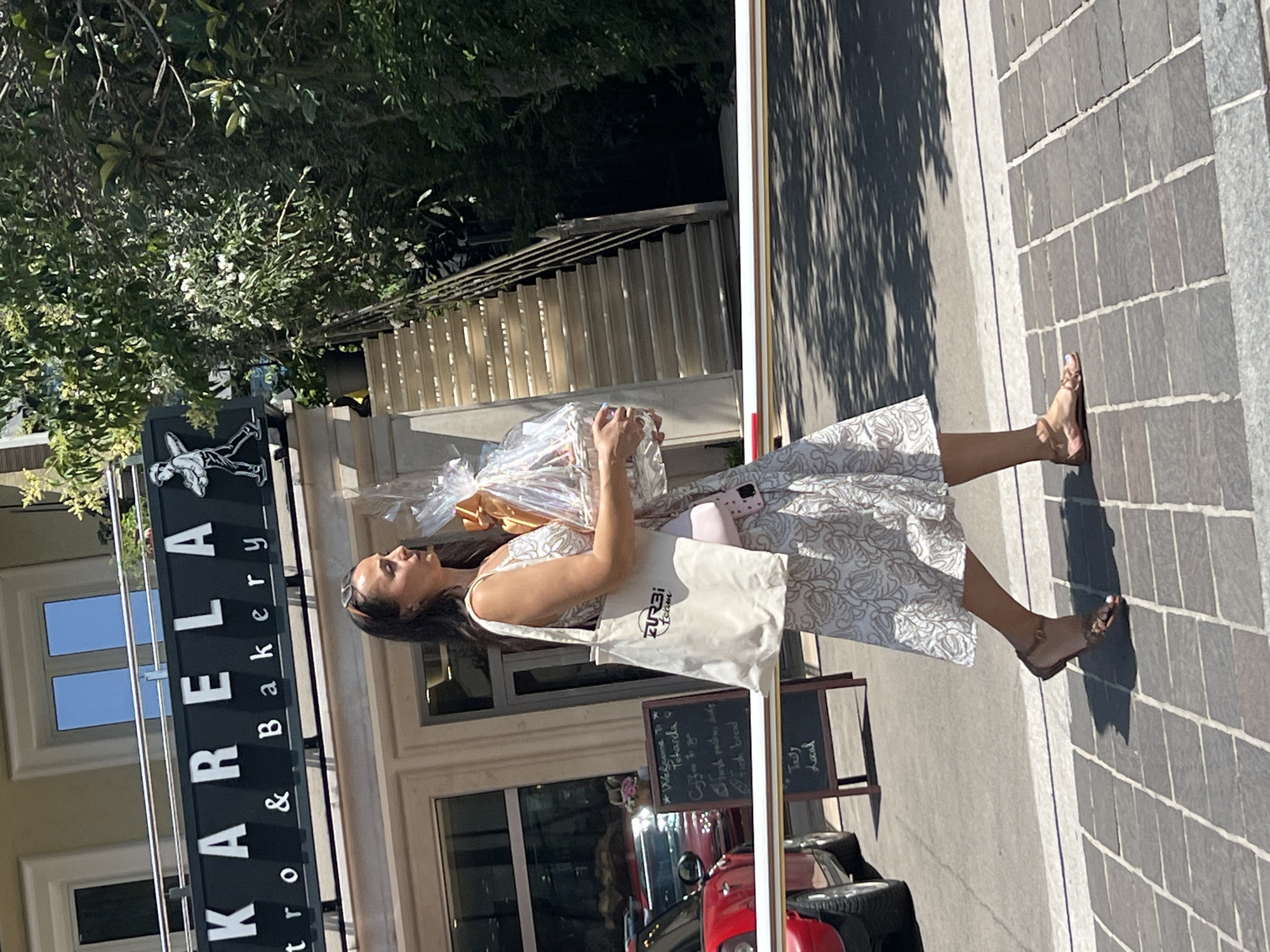 A woman walks past the "KARELA Bistro & Bakery" in Budva, Montenegro, carrying a basket of food. She is wearing a white patterned dress and brown sandals. The bakery has a black sign with white lettering and a small graphic of a person holding a tray. To the left of the bakery, a red car is parked. A menu board is visible on the sidewalk in front of the bakery. Behind the woman, a set of outdoor stairs leads upwards, flanked by lush green trees and foliage. A white barrier is positioned across the path in the foreground. The ground is paved with dark square tiles.
