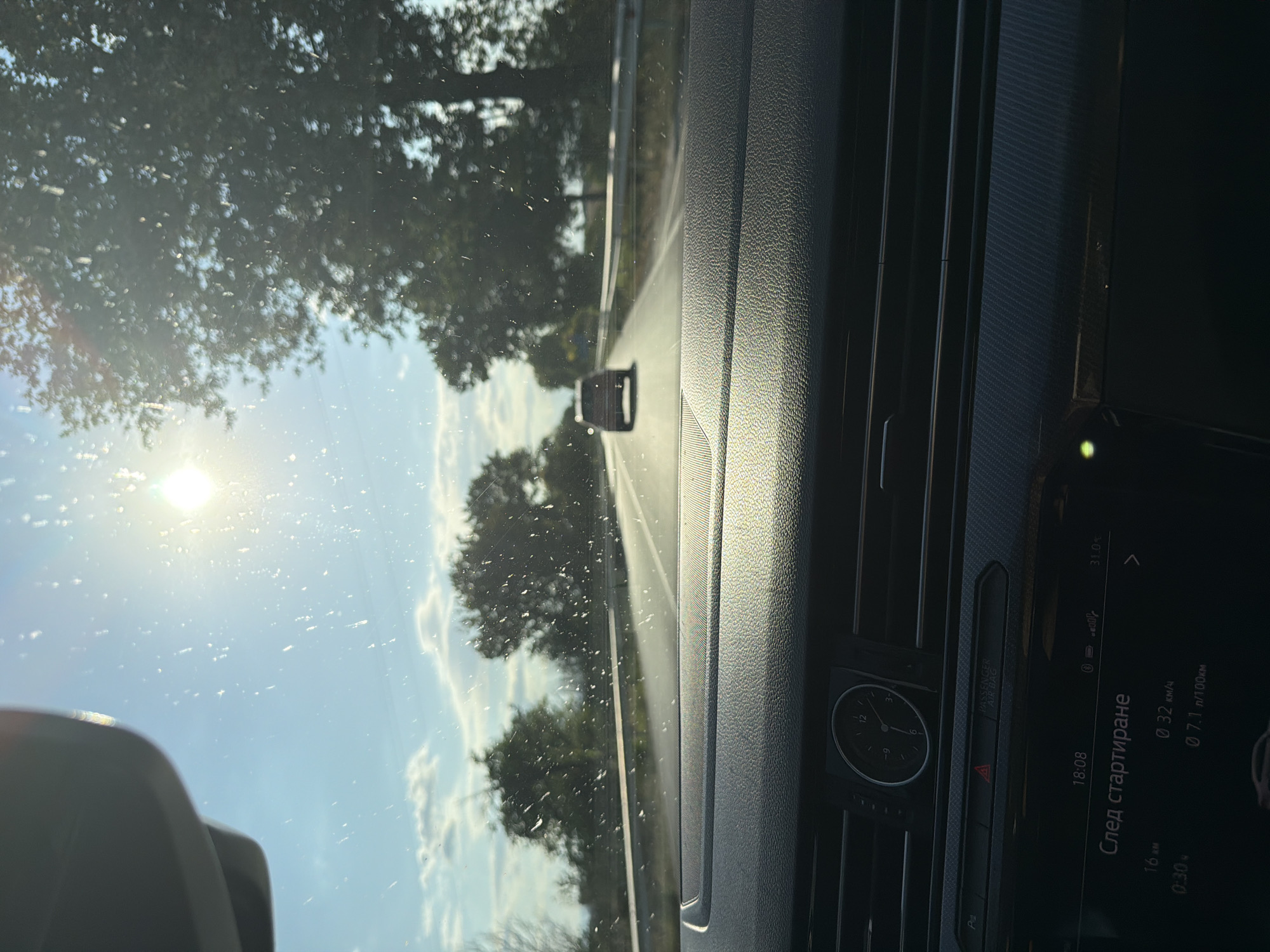 The image is taken from inside a car, looking forward through the windshield. The sun is shining brightly, creating lens flare and glare on the glass. The road ahead is clear, with another car visible in the distance. Trees line the sides of the road, and the sky is blue with some clouds. The interior of the car is visible in the foreground, including the dashboard and a clock. The display on the dashboard shows the time as 18:08 and other information such as distance traveled, speed, and fuel consumption. The scene appears to be a sunny day on a highway, possibly in a rural area.