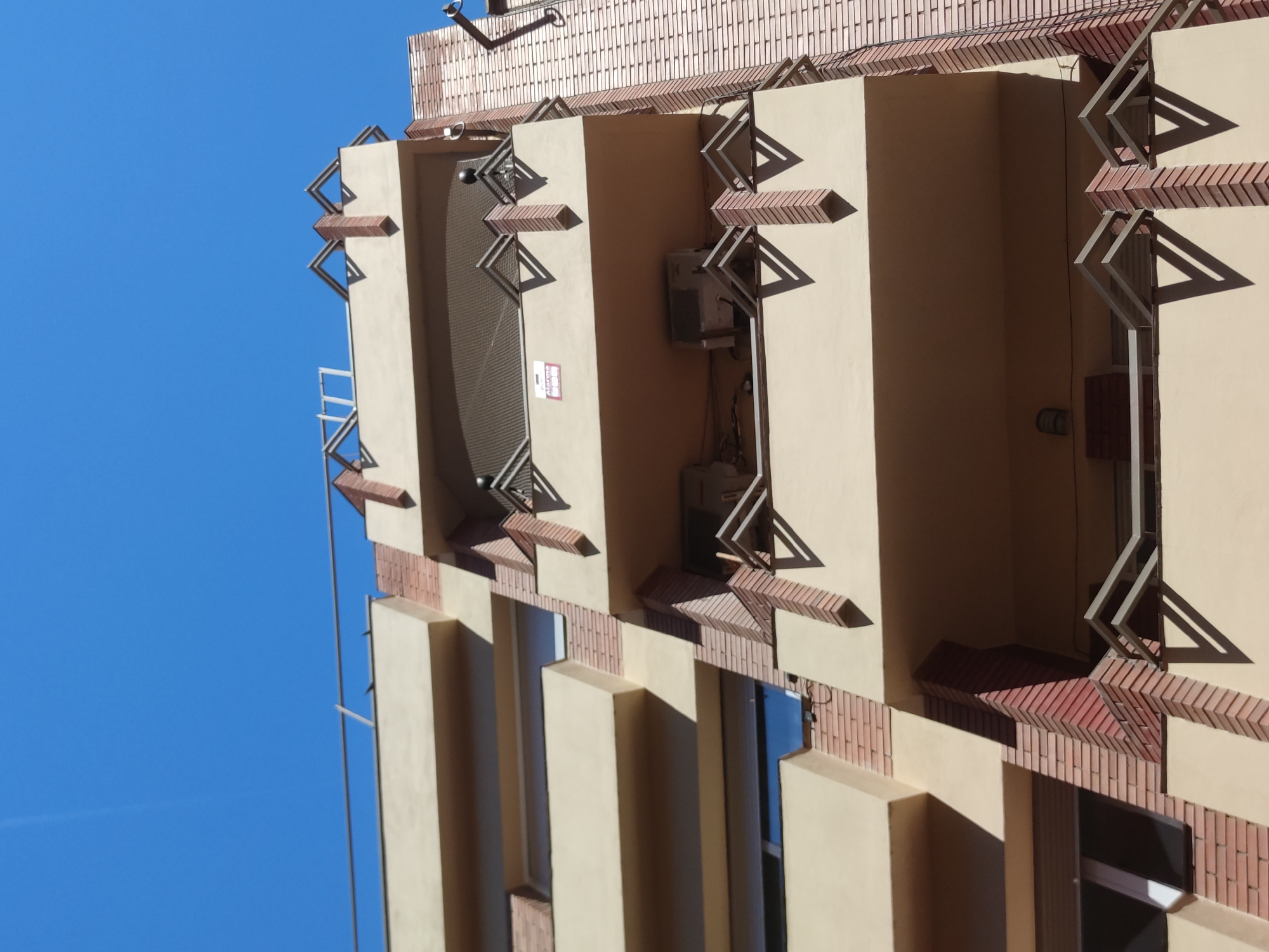 This image captures a portion of a building's facade, with its architectural features prominently displayed against a clear blue sky. The building has a light-yellow or beige hue and is accented with brickwork in reddish-brown tones. The design incorporates rectangular protrusions that create a segmented or layered effect. Notably, the structure features decorative elements resembling open, triangular frames, possibly made of metal or another light-colored material.
An air conditioning unit is visible between the architectural protrusions. The presence of windows and metal fixtures indicates the building's functional aspect. The image does not include any people or other distinct subjects. The composition is focused on the architecture and the interplay of light and shadow.