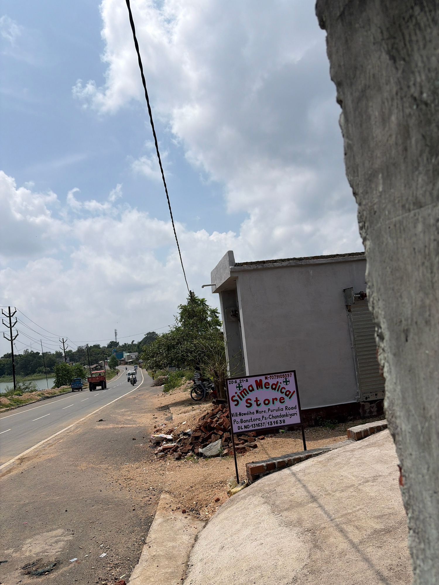 The image shows a road scene in Banshtora, India, with a small building and a sign for "Sima Medical Store." The sign is black with white and pink text, and it provides the address "Vill-Nowdiha More, Purulia Road, Po:-Banstora, Ps:-Chandankiyari" along with a DL number.
The road is asphalt, with white lane markings visible. There is a body of water to the left of the road, likely a river or lake, with trees and vegetation along its bank. Various vehicles are present on the road, including a red tractor, a blue truck, a white van, and motorcycles. Power lines and poles run along the side of the road.
The building in the foreground, next to the medical store sign, is a light gray, rectangular structure with a roll-up metal door. To the right of the image, a rough, textured gray wall is partially obscuring the view. The sky is partly cloudy, with a mix of blue sky and white clouds. There is a pile of bricks near the medical store sign, and some litter is scattered on the roadside. The overall impression is a somewhat rural or semi-urban setting in India.