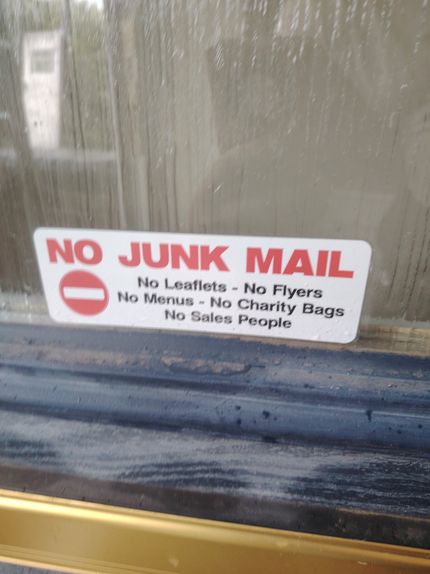 A sticker with bold red lettering reads "NO JUNK MAIL". Below this, in smaller black text, are the words "No Leaflets - No Flyers", "No Menus - No Charity Bags", and "No Sales People". A red and white prohibition symbol is to the left of the "NO JUNK MAIL" text. The sticker is affixed to a glass surface, possibly a door or window, which appears to be wet, with water droplets visible. Outside the glass, a blurred background suggests a building or structure with vertical lines, possibly due to rain streaks or condensation. The bottom of the image shows a dark blueish-grey surface, possibly a window frame or trim, with a golden-colored edge beneath it. The scene is likely in a residential or commercial area, possibly a doorstep in Huntingdonshire, United Kingdom, given the context of junk mail and sales people.