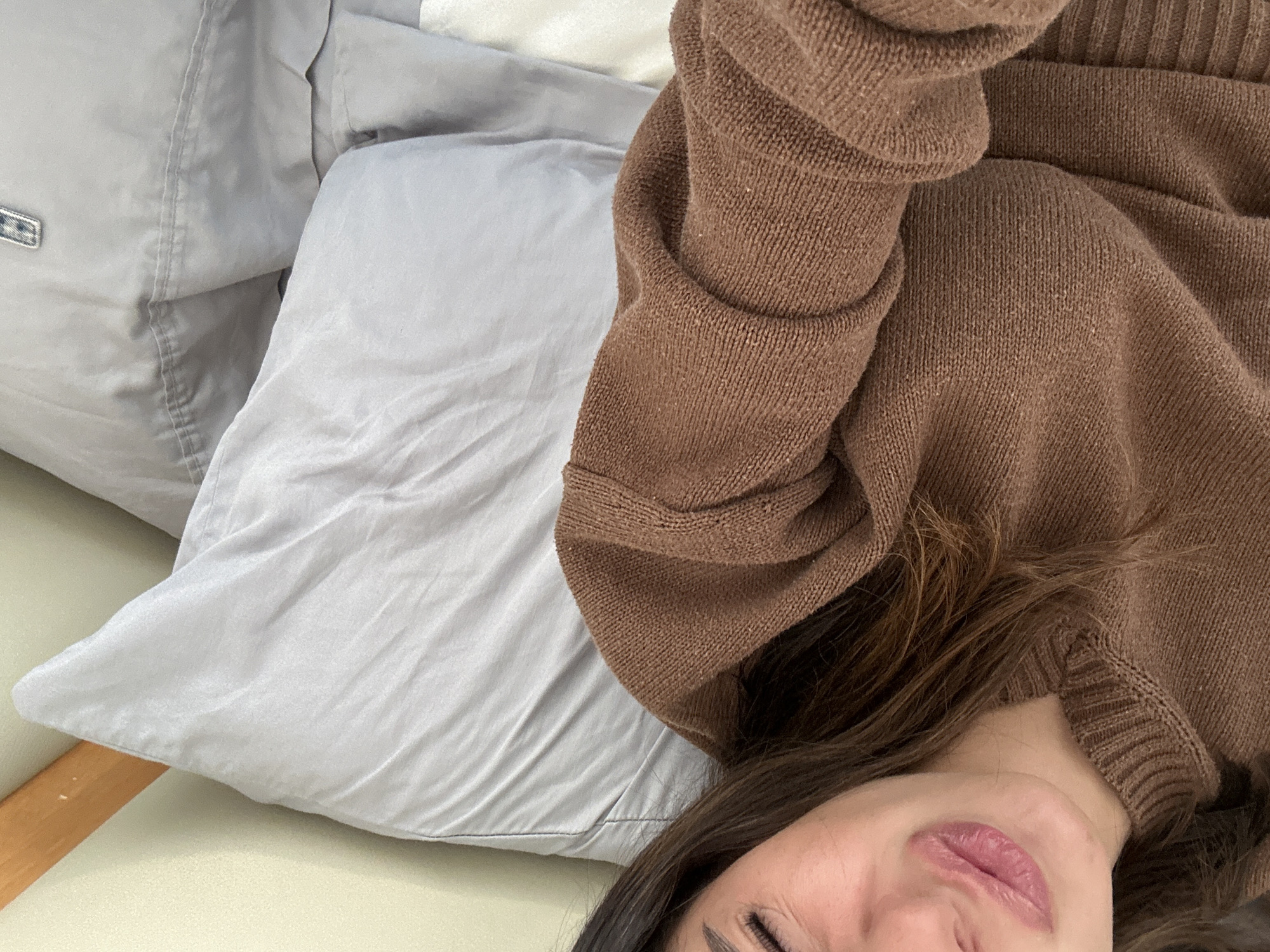Here is a description of the image:
A woman is lying on a bed or couch with grey pillows. She is wearing a brown sweater. The location is unknown.