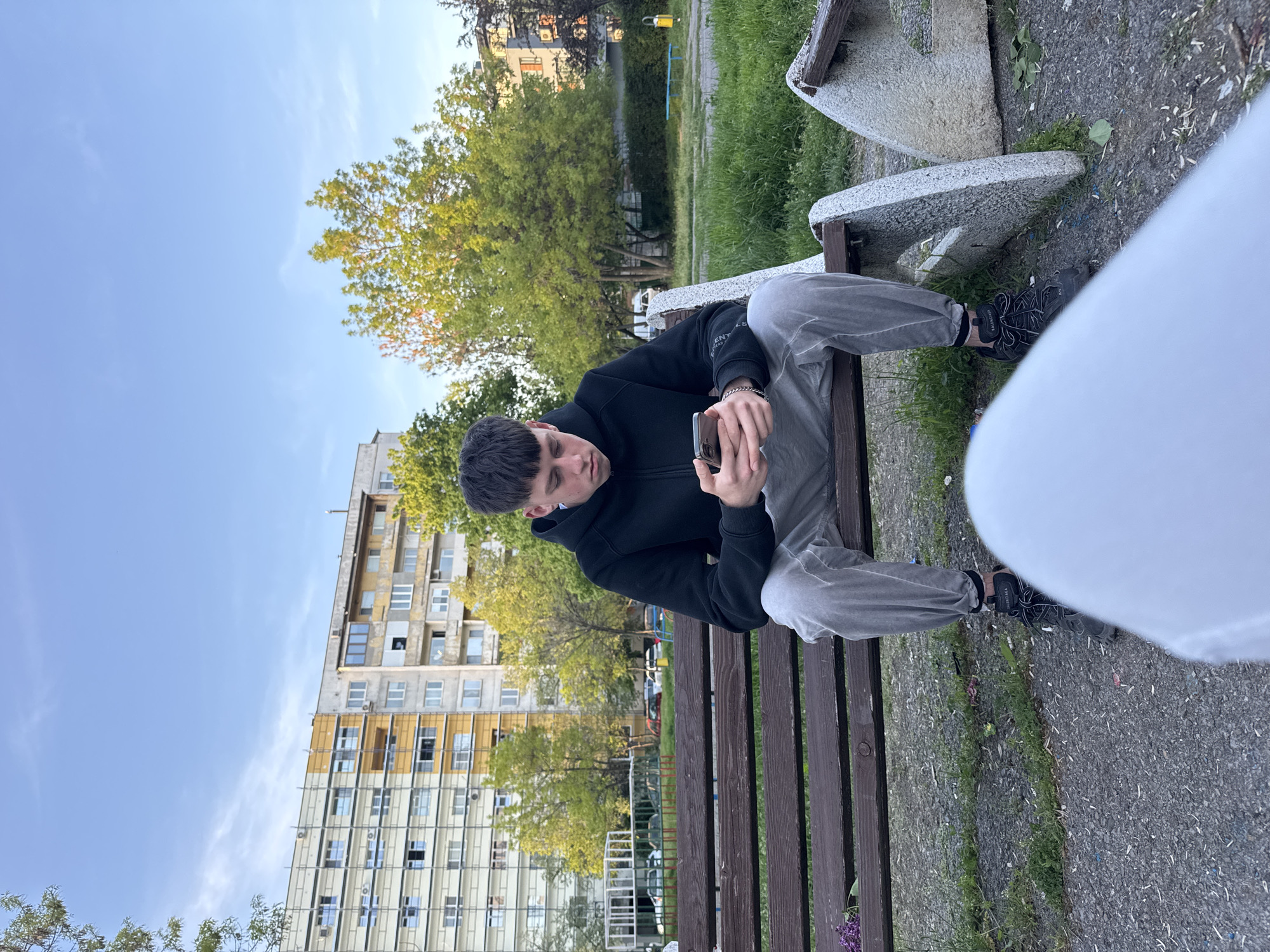 A young man is seated on a wooden park bench with concrete supports, engrossed in his smartphone. He is positioned slightly off-center to the left, looking down at the device held in both hands. He wears a black zipped hoodie with a partially visible white text "EN ES" on the sleeve, light grey baggy pants, and black sneakers where the brand "BALENCIAGA" is discernible on the right shoe. A silver chain bracelet is visible on his left wrist.
The setting appears to be a public park or green space within an urban environment. In the mid-ground, behind the man, are several trees with vibrant green foliage, illuminated by natural light suggesting late afternoon or early evening. Beyond the trees, a large, multi-story apartment building with numerous windows and balconies dominates the background. Sections of the building feature an older, somewhat weathered facade, while other parts are updated with yellowish panels. The sky above is clear and blue with very few faint clouds.
The ground around the bench is a paved surface, likely asphalt or gravel, with patches of green grass and small weeds. A large, blurred white object occupies the right foreground, partially obscuring the man's right leg and foot, and the ground directly in front of him. The overall scene depicts a casual moment of an individual interacting with technology in an outdoor, community setting, consistent with an urban park in a place like Sofia, Bulgaria.