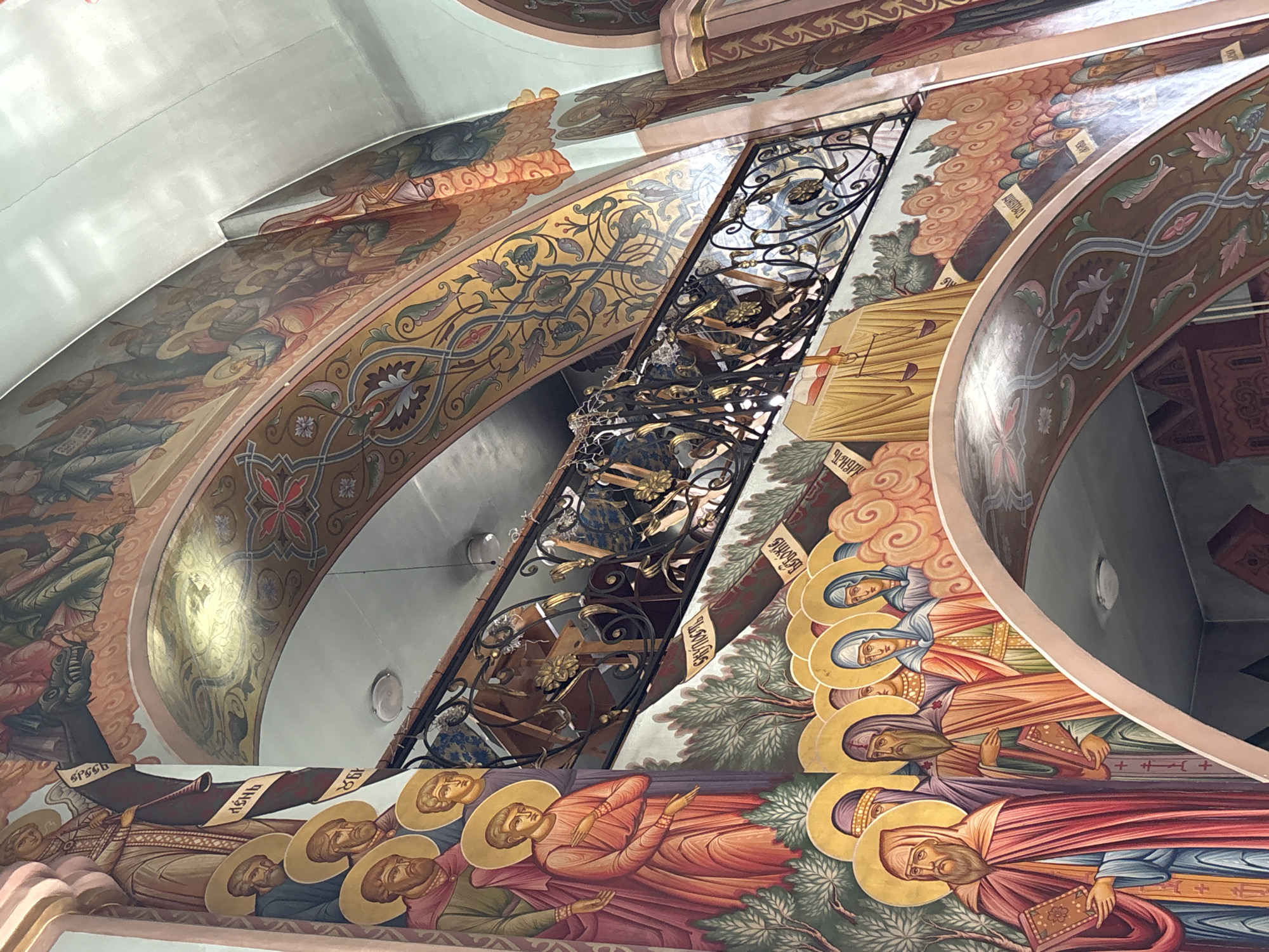 The image shows an interior view of a church in the city of Chaikovsky, Russia. The scene is dominated by ornate religious murals depicting biblical figures and scenes painted on curved arches and ceilings. There are several saints and other religious figures. A black, wrought-iron chandelier hangs from the ceiling.