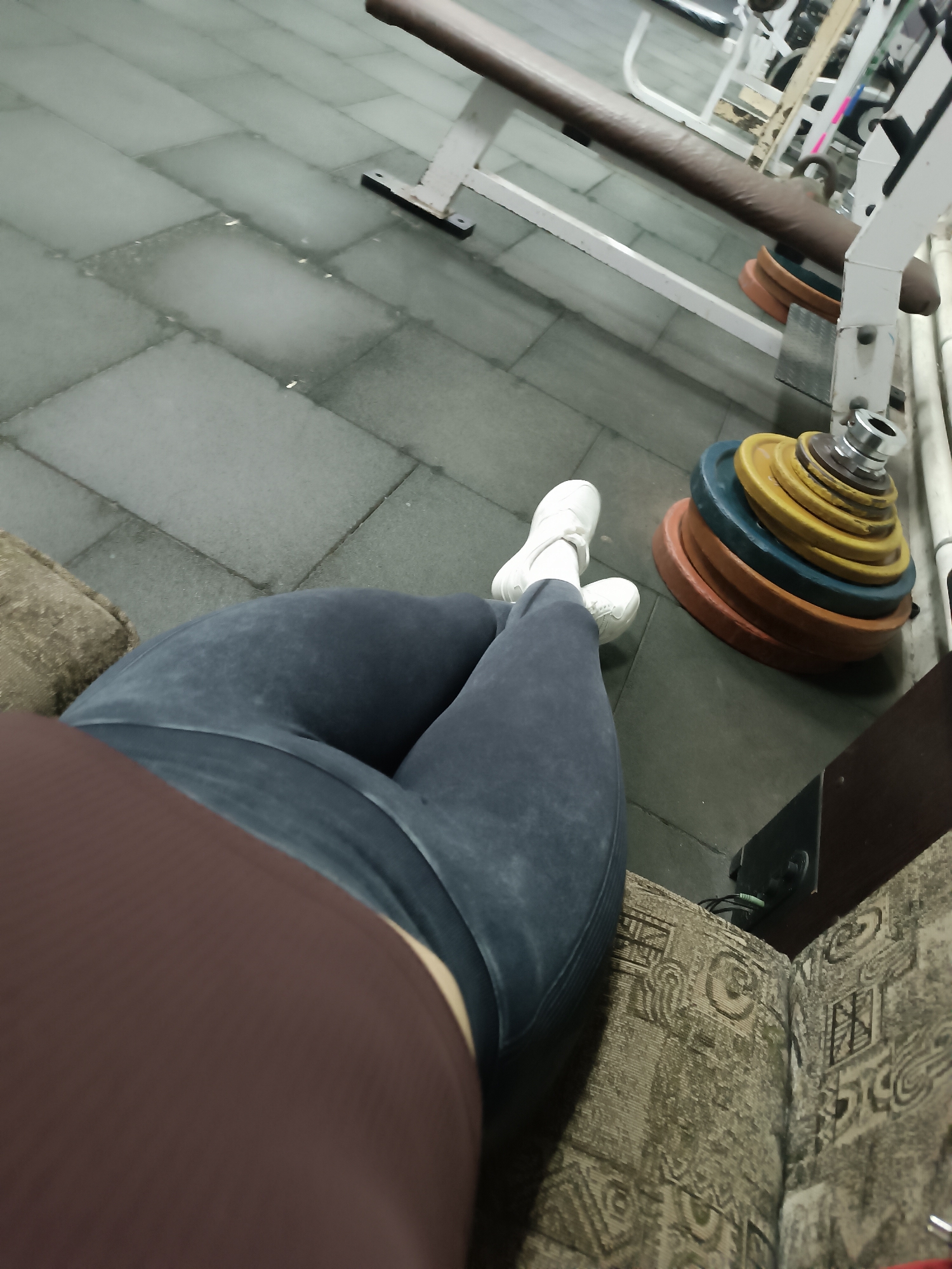The image shows a person sitting on a patterned couch. The person is wearing a brown shirt and blue leggings, with white sneakers on their feet. The location appears to be a gym, with weights stacked on the floor. There's also workout equipment visible in the background. The city is unknown.