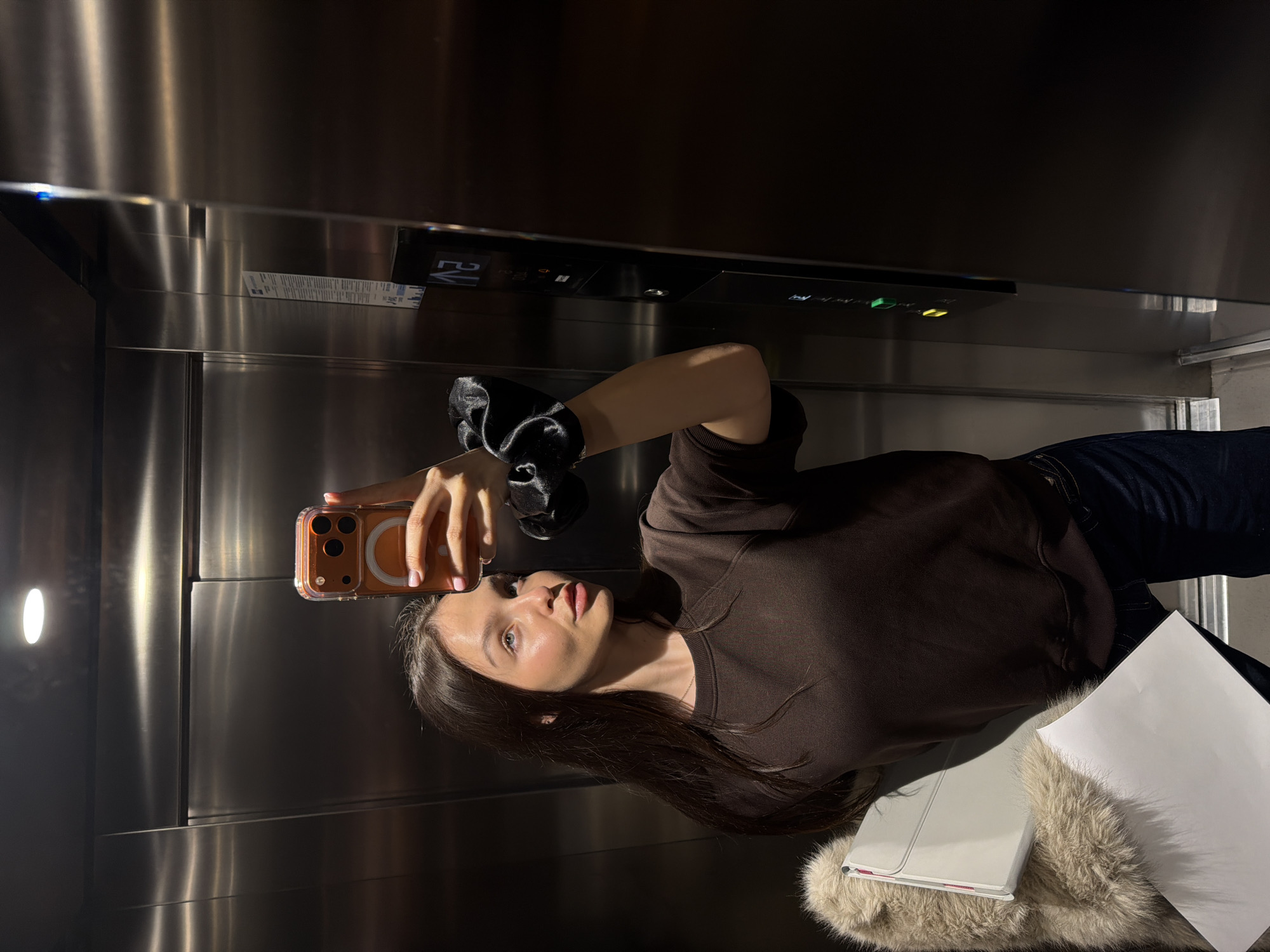 A young adult female is depicted taking a selfie photograph inside an elevator in Sofia, Bulgaria. The elevator interior features polished stainless steel walls and a circular overhead light fixture. The female subject has long dark hair and is wearing a dark brown sweatshirt and dark blue jeans. She holds an orange-cased Apple iPhone with her left hand, positioned to capture her reflection in the polished surface. A black satin scrunchie is visible on her right wrist. Under her left arm, she holds a light beige fur-lined item and two white rectangular objects, identified as a folder and loose papers. To the right, an elevator control panel is visible, displaying "Λ2" (indicating floor 2 with an upward arrow) and illuminated green and yellow buttons. A vertical informational sticker from 'Technolift OOD' on 'ul. Kozloduy' (Kozloduy Street) in Sofia is affixed to the elevator wall, confirming the location.