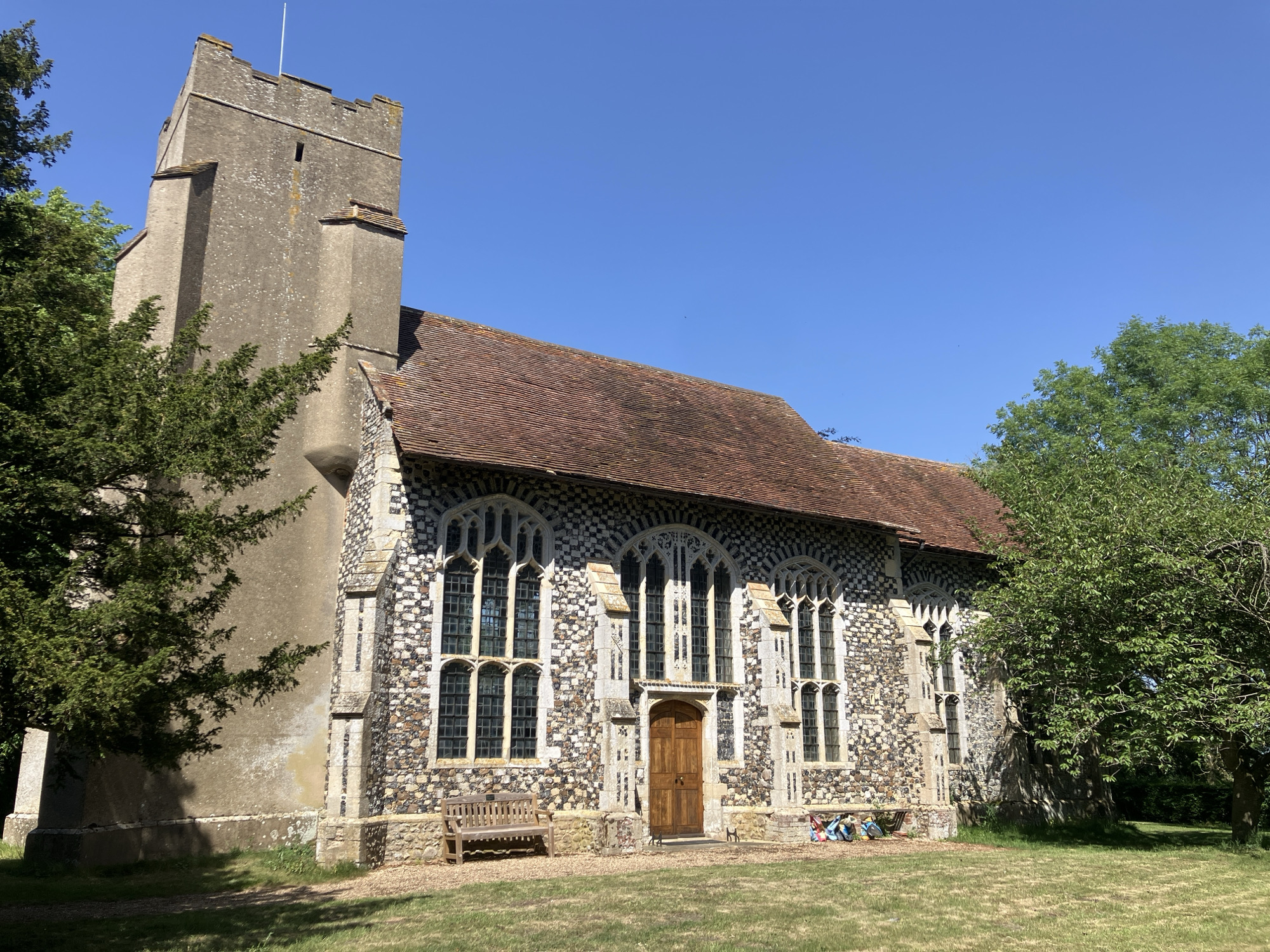 The image depicts St Peter's Church in Brome, Mid Suffolk, United Kingdom, a stone building with a tall tower and a tiled roof. The church has multiple arched windows, and a wooden bench sits in front of it on a lawn of dry grass. A wooden door is set in the middle of the front wall of the church. Trees partially obscure the building on both sides, and the sky is clear and blue.