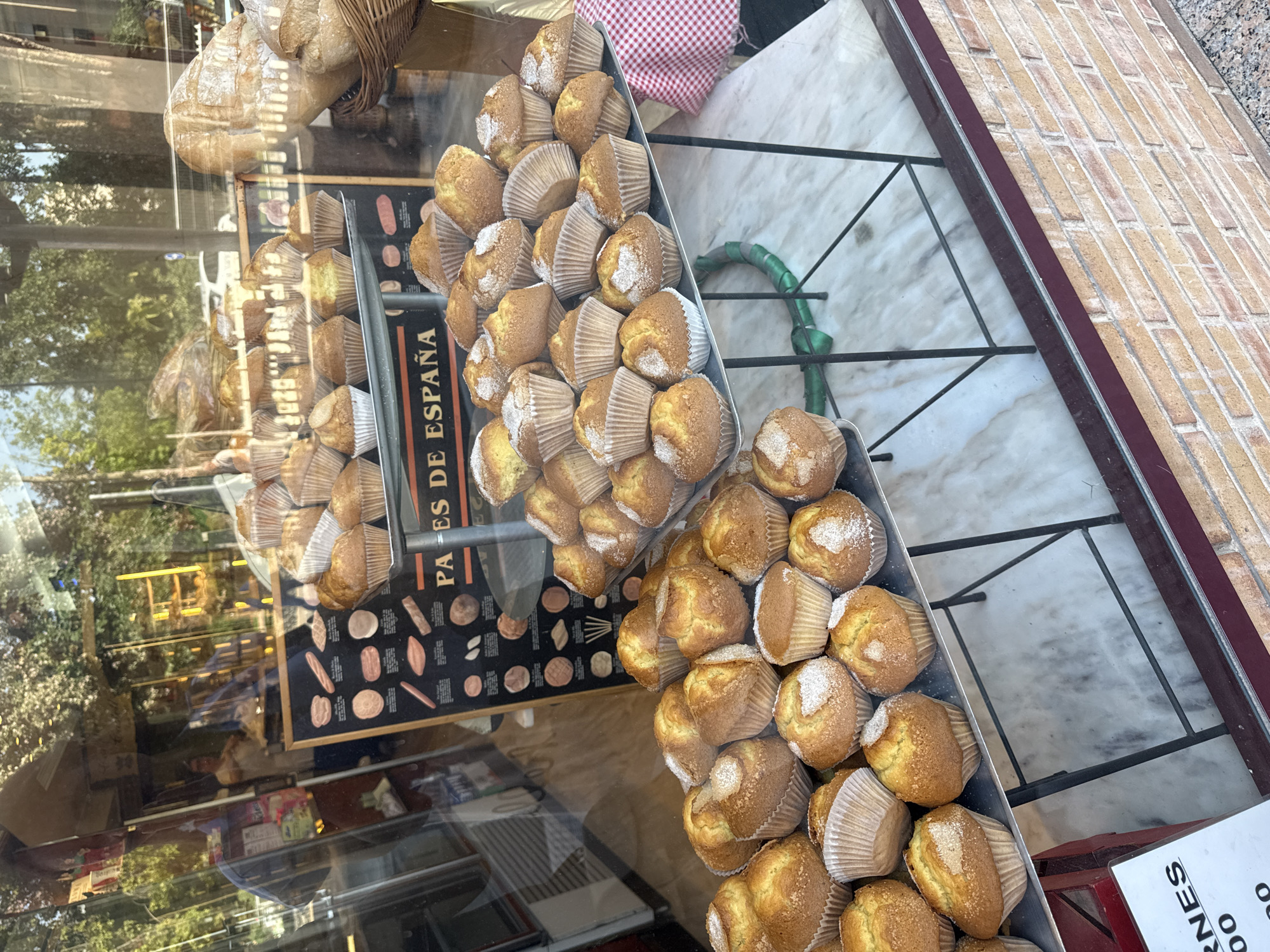 The image captures a close-up view of a bakery display window, showcasing an abundance of baked goods, primarily muffins or cupcakes, arranged on metal trays. These light-colored pastries are set in white paper liners and are often topped with a dusting of granulated sugar, giving them a glistening appearance. Multiple trays are stacked or placed behind the glass, with more baked items, possibly loaves of bread, visible further back and higher up in the display.
Reflections on the glass obscure some parts of the interior and overlay the view with an outdoor scene. These reflections show green foliage from trees, a glimpse of what appears to be a street or outdoor area, and other indistinct elements from the environment outside the shop. A checkered red and white fabric is visible behind one of the trays, and the display area features a mix of surfaces, including a white marble-patterned countertop and a darker surface with a grid-like pattern, possibly a metal stand. The exterior of the shop features a brick-patterned wall section.
A prominent black sign with white illustrations of various bread shapes is visible through the glass. Text on this sign reads "PAES DE ESPAÑA," suggesting "Breads of Spain" or "Spanish Breads," indicating the location is likely in a Spanish-speaking region. Another white sign in the bottom right corner shows partially visible text "RNES" and numbers "00," which might be part of a price list or product description. The lighting suggests it is daytime, with bright natural light illuminating the scene. The overall impression is a vibrant display of fresh baked goods in a bustling urban environment.