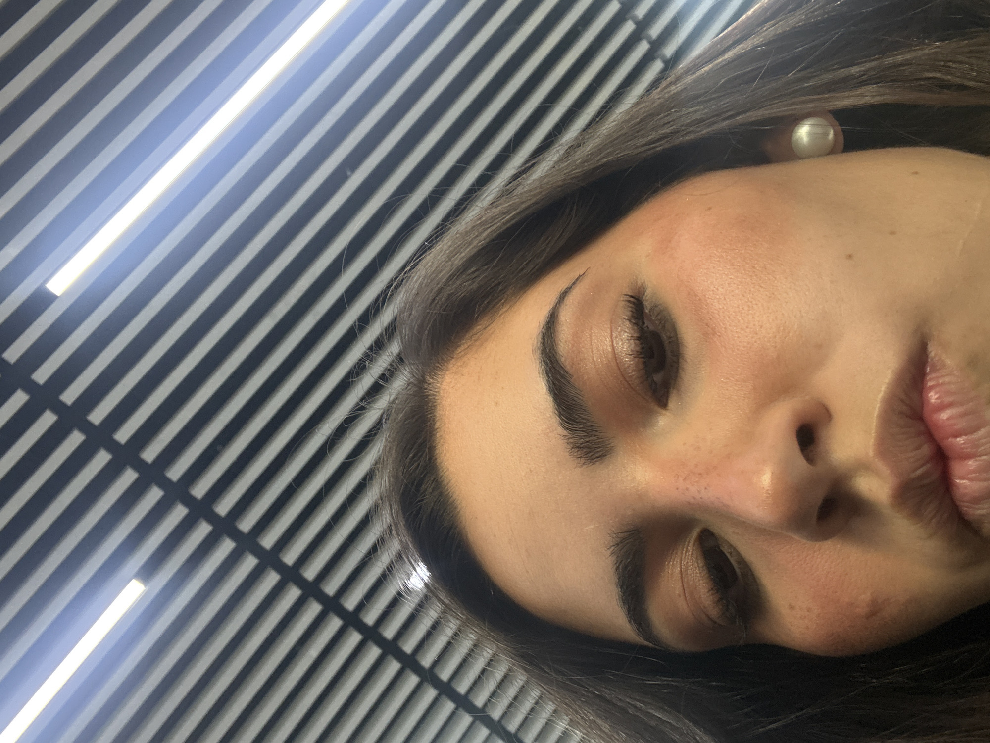 A close-up, angled portrait of a woman with dark hair and fair skin. She has prominent eyebrows and closed eyes. The background features a pattern of alternating light and dark gray stripes, suggesting a ceiling with parallel light fixtures. A round, light-colored earring is visible on the woman's ear. The lighting is bright and somewhat harsh. The image does not contain information indicating a location in Mexico City, Mexico.