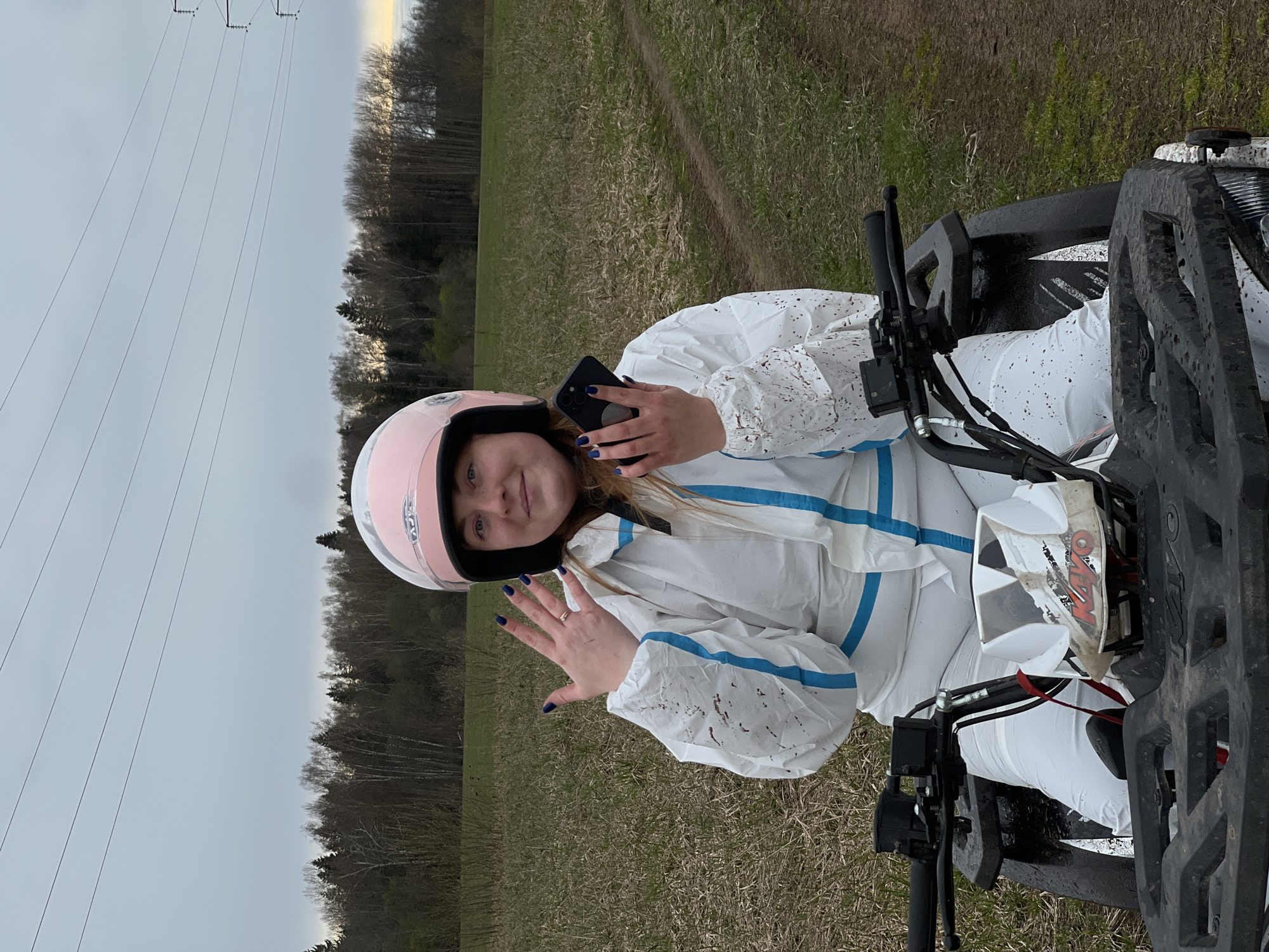 A person wearing a pink helmet and a white protective suit with blue stripes is sitting on an all-terrain vehicle (ATV) in a field. The person is holding a smartphone in their left hand and waving with their right hand. There are trees and power lines visible in the background. The location is likely in the Razdolye area, Russia, given the prompt. The field appears to be grassy and slightly uneven. The ATV is partially visible, with handlebars and front sections evident. The person's clothing has some dirt spots on it. The sky is overcast.