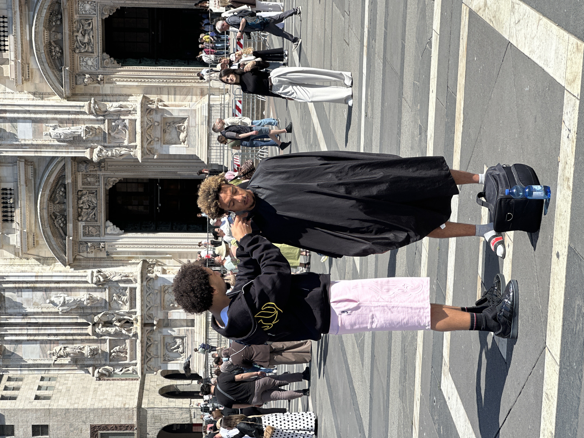 Two men are standing in front of the Duomo di Milano in Milan, Italy. One man is wearing a black jacket and pink shorts. The other man is wearing a black robe. There are many people walking around in the background.