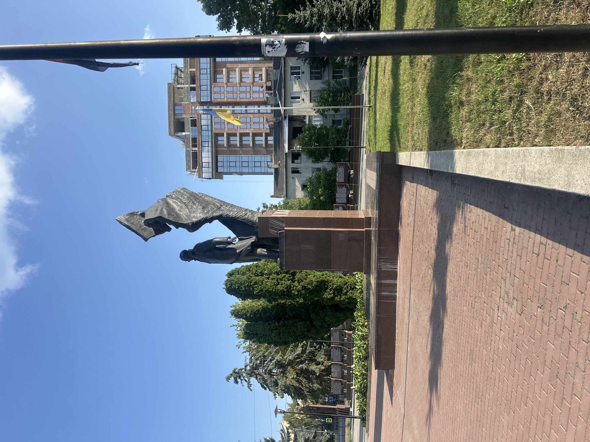 The image depicts a monument in a public square, likely in Ternopil, Ukraine, given the presence of the Ukrainian flag. The monument itself is made of dark metal and depicts two figures, one standing and one appearing to be in motion or part of a larger structure. The monument is situated on a tiered, dark-colored base. In the background, a modern building with glass windows and visible construction elements is under development. To the left of the monument, there are trees, including coniferous and deciduous varieties, and a street lamp. A Ukrainian flag is prominently displayed on a flagpole in the mid-ground. The foreground features a red brick paved area, and to the right, a patch of green grass with some trees. The sky is blue with scattered clouds.