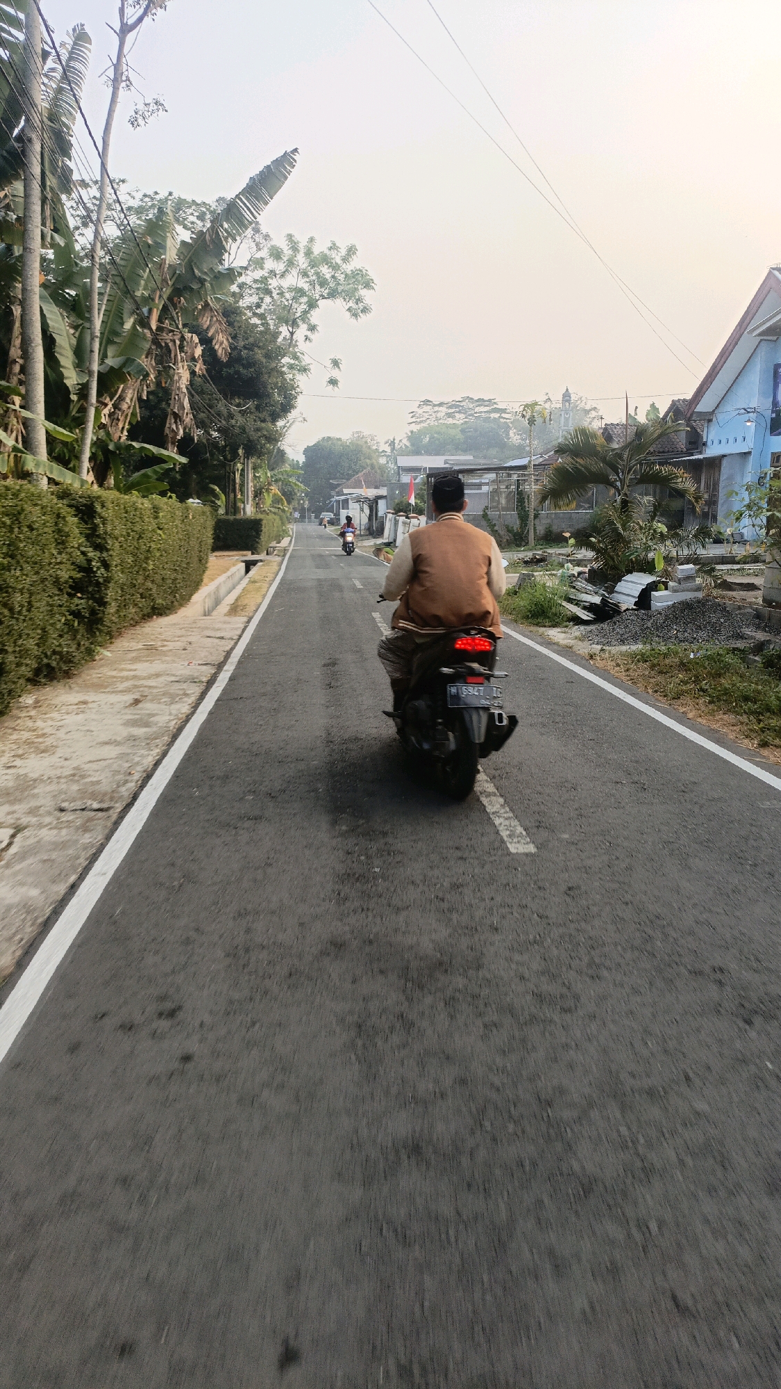 A medium shot captures a person riding a black scooter from behind on a straight, paved road. The rider, wearing a brown jacket and a black cap, is moving away from the camera. The road is bordered by a white line on the left and has scattered dark patches. To the left of the road, a neatly trimmed green hedge runs alongside a concrete path. In the distance, more people on scooters are visible, and the scene is framed by trees and buildings. The sky is overcast with a light haze. This road appears to be located in a residential area of Salatiga, Indonesia, possibly on the outskirts of the city, given the presence of vegetation and modest houses.