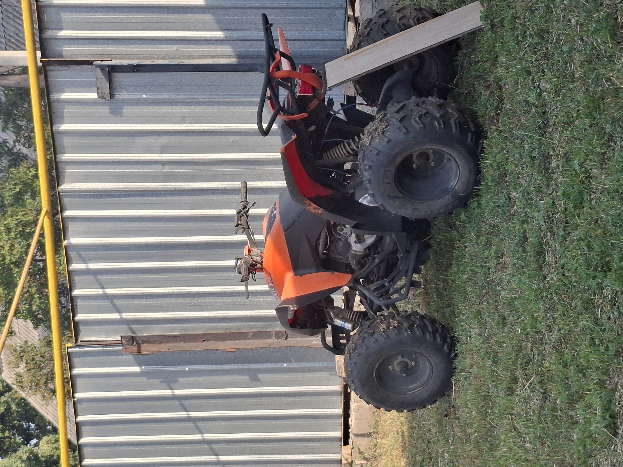 A red and black quad bike is parked on a grassy area in front of a corrugated metal fence. A yellow pipe runs horizontally above the fence, and trees and a roof are visible in the background. The quad bike has large, knobby tires and a luggage rack at the rear. A piece of light-colored wood leans against the rear of the quad bike. The scene appears to be outdoors, possibly in a rural or suburban area of Almaty, Kazakhstan. There are no people visible in the image.