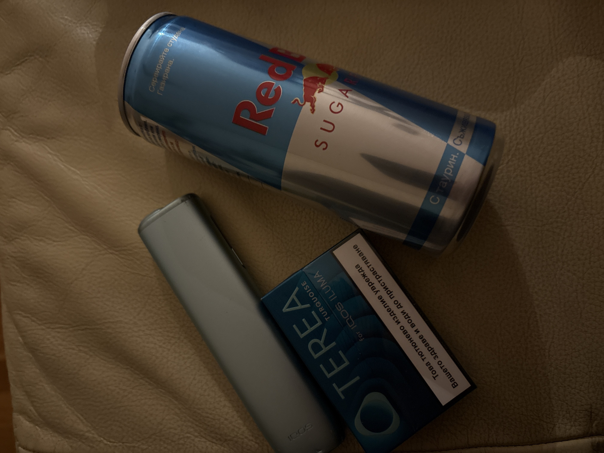 On a tan, possibly leather, surface are a Red Bull energy drink can, an IQOS Iluma electronic cigarette device, and a pack of Terea Turquoise for IQOS Iluma tobacco sticks. The energy drink can is predominantly blue and silver with red lettering and the Red Bull logo. The electronic cigarette device is light grey, and the Terea pack is a dark blue with a white warning label written in Bulgarian, advising that the product harms health and leads to addiction.