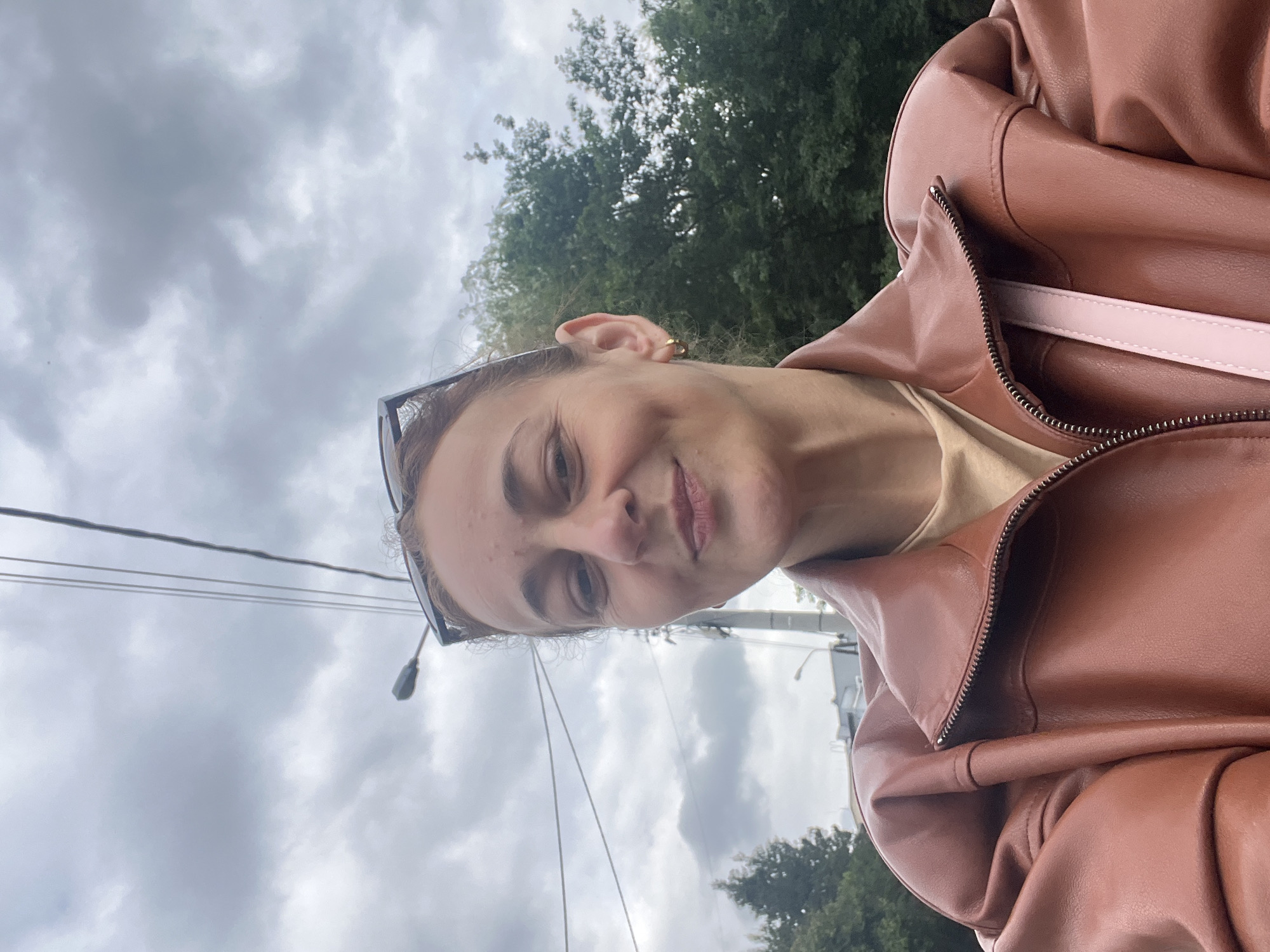 A woman is taking a selfie outdoors on a cloudy day. She is wearing a brown leather jacket and a light-colored top. Her hair is pulled back, and she is wearing sunglasses on her head. She has a dimple on her left cheek and is wearing a hoop earring in her left ear. The background shows trees, a utility pole with wires, and some buildings in the distance. The setting appears to be a street or park in an urban area.