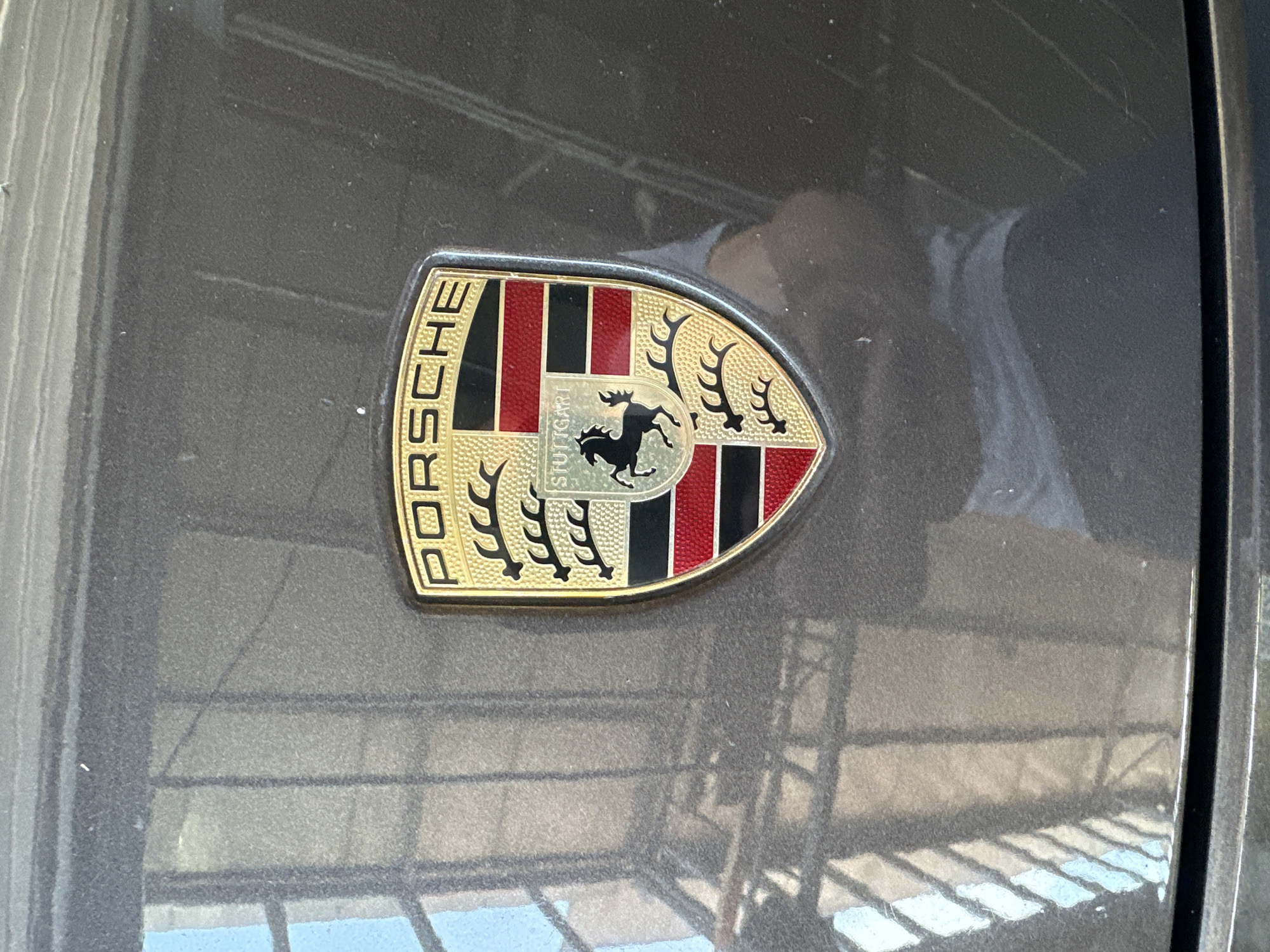 The image shows a close-up of a Porsche emblem on a dark gray car. The emblem is a crest with the word "PORSCHE" written in gold letters along the top edge. The crest is divided into sections, featuring red and black stripes, a black horse in the center, and antlers on either side. The word "STUTTGART" is also visible below the horse. The car's surface appears to have some dust and reflections, suggesting it is outdoors or in a garage. The reflections on the car's surface show a grid-like pattern, possibly from a ceiling or structure above, and some bright rectangular shapes that could be windows or light sources. The lighting suggests it is daytime. There are no people visible in the frame.