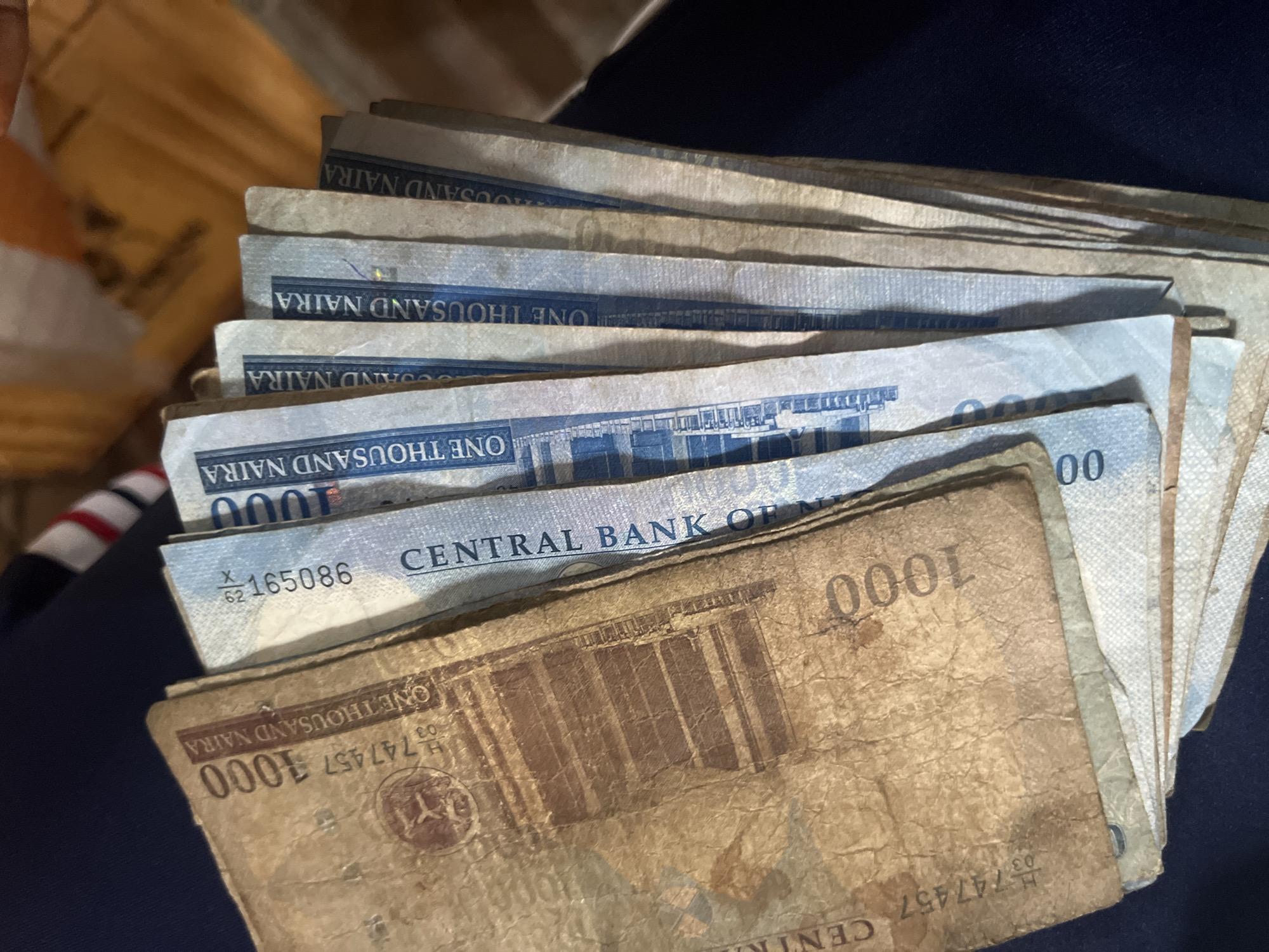 The image displays a fanned arrangement of Nigerian Naira banknotes, primarily the 1000 denomination. The bills are in varying conditions, with one prominent bill in the foreground appearing aged and worn, exhibiting a sepia tone and visible creases. This bill has the number "1000" clearly printed on it, along with "ONE THOUSAND NAIRA" and a serial number starting with "H 747457". Behind this, a stack of newer-looking blue and white 1000 Naira notes is visible, with "ONE THOUSAND NAIRA" and "CENTRAL BANK OF NIGERIA" printed on them. Some of these newer bills have serial numbers such as "X 62 165086". The banknotes are spread out over a dark blue fabric surface, which appears to be clothing. The background is out of focus but suggests an indoor setting with warm lighting, possibly a desk or table with some yellow-tinted items. The photograph was taken at an angle, looking down at the bills. There is no specific indication of the city of Nkpor, Nigeria within the image itself.
