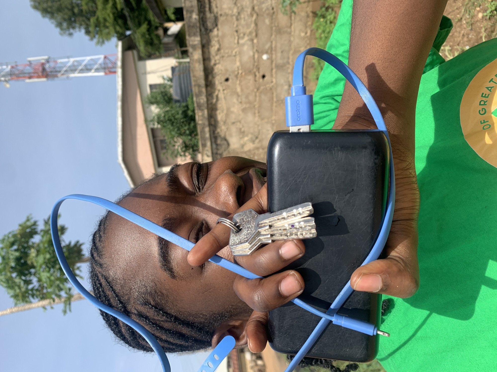 The image captures a close-up, outdoor shot of a person in Rafin Par, Nigeria, presented on its side. The individual, who has dark skin and braided hair, appears to be a young adult, with eyes closed or squinting, possibly due to bright sunlight, and a subtle smile. They are wearing a bright green t-shirt with a partial logo visible that reads "...OF GREAT...".
In their hand, the person holds a black, rectangular device, likely a power bank, and a set of silver keys on a metal ring. A bright blue cable is prominent, with one end, a USB-A connector bearing the brand "arafmo", plugged into the black device. The other end of the cable appears to be a 3.5mm audio jack, visible near the person's ear/shoulder.
The background shows a clear, bright blue sky and an outdoor setting. Visible elements include a tall communication tower, a brick wall, part of a building, and green foliage from trees and bushes. The strong shadows suggest direct sunlight, indicating a sunny day, likely midday or early afternoon.
