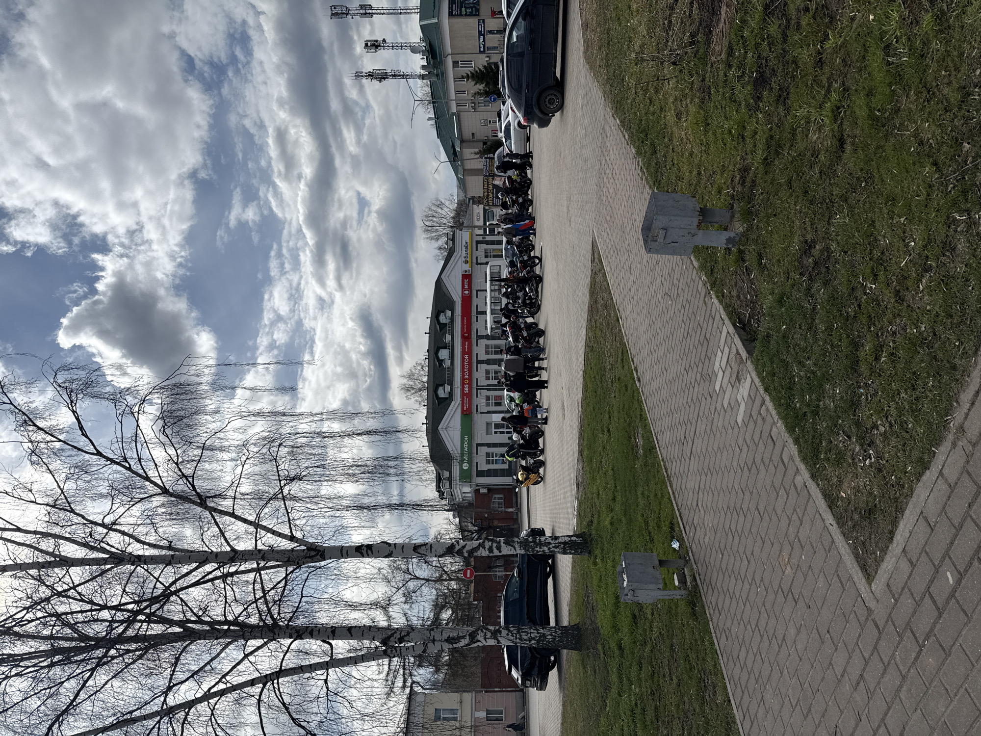A street scene in Glazov, Russia, shows a row of motorcycles parked along a brick walkway adjacent to a grassy area. A building with a prominent "Мегафон" sign and another with "585 Золотой" are visible. A row of bare trees is in the foreground, and overcast skies dominate the background. A car is parked nearby. The scene appears to be a gathering of motorcyclists. Cellular towers are also observed in the distance.