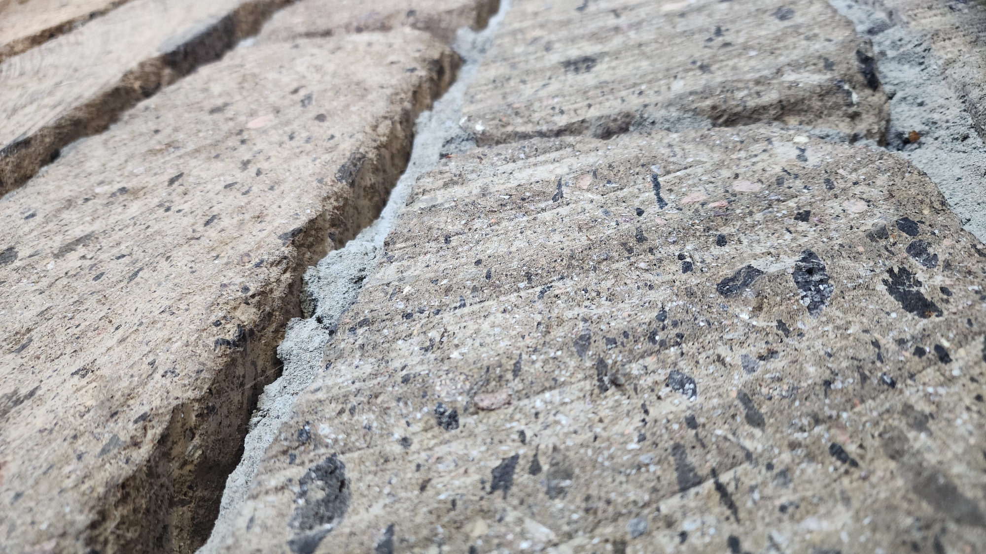 The image is a close-up, macro photograph of a brick wall, focusing on the textured surface and mortar. The bricks are a light brown or tan color, with visible aggregate and small dark inclusions. The mortar is a lighter grey and appears somewhat rough and uneven. The composition is angled, showing the texture of the bricks and the way they are joined together. There are no people, actions, or interactions depicted in the image. The scene is static and focuses solely on the material and construction of the wall. The location in Azatavan, Armenia, is not visually represented within the image itself; it serves only as contextual information.