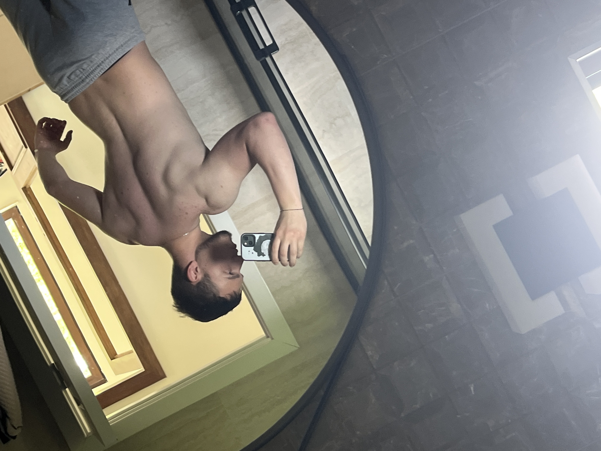 In this mirror selfie taken in Войводино, Bulgaria, a young man is captured from the waist up. He is shirtless, wearing grey shorts, and displays a muscular physique, particularly in his back, shoulders, and arms. He has short dark hair and a beard. The man holds a smartphone in his right hand; the phone features a light-colored case with a black graphic, possibly a stylized animal or character. A thin chain is visible around his neck, and a subtle bracelet on his left wrist.
The large, circular or oval mirror reflects an indoor setting, likely a bathroom, illuminated by bright daylight. To the left within the reflection, a wooden-framed window or doorway allows significant natural light to enter, suggesting the photo was taken during the day. The walls appear light-colored, possibly cream or beige. On the right side of the reflection, dark tiled walls are visible, partially obscured by a bright lens flare originating from a light source or window. A dark horizontal bar, potentially part of a shower enclosure or fixture, is also visible above the reflection of the man's head. The overall impression is a personal moment captured in a well-lit space.
