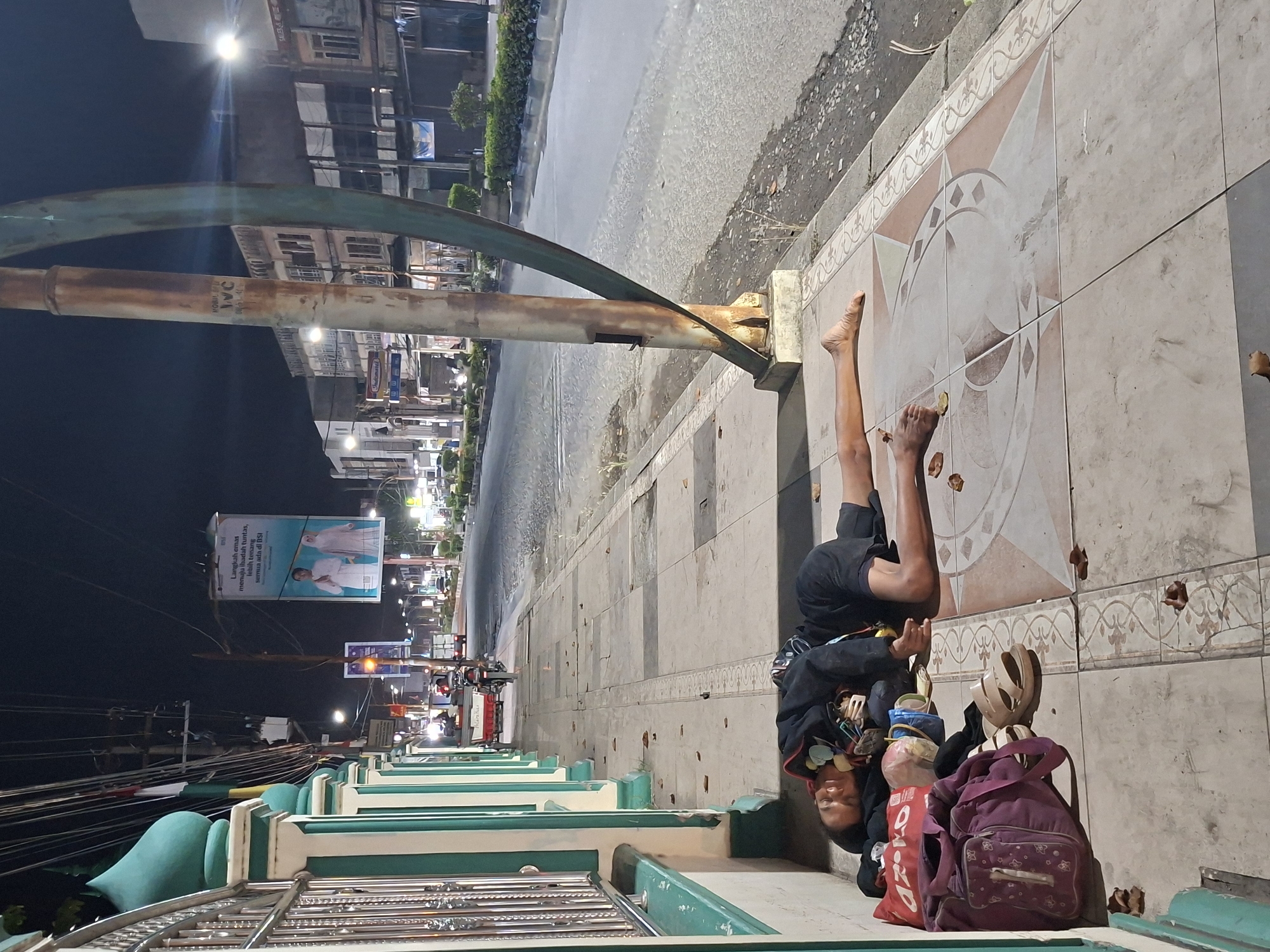 A person in black shorts and a jacket lies on their back on a tiled sidewalk in Lhokseumawe, Indonesia. Their bare feet point towards a street with a wet surface and streetlights. A metal pole spans over the sidewalk, originating from the ground near the person's feet. The street is lined with buildings. A purple backpack, a red bag, and other items are next to the person. A green and white structure is in the foreground, and a large banner is visible in the distance.