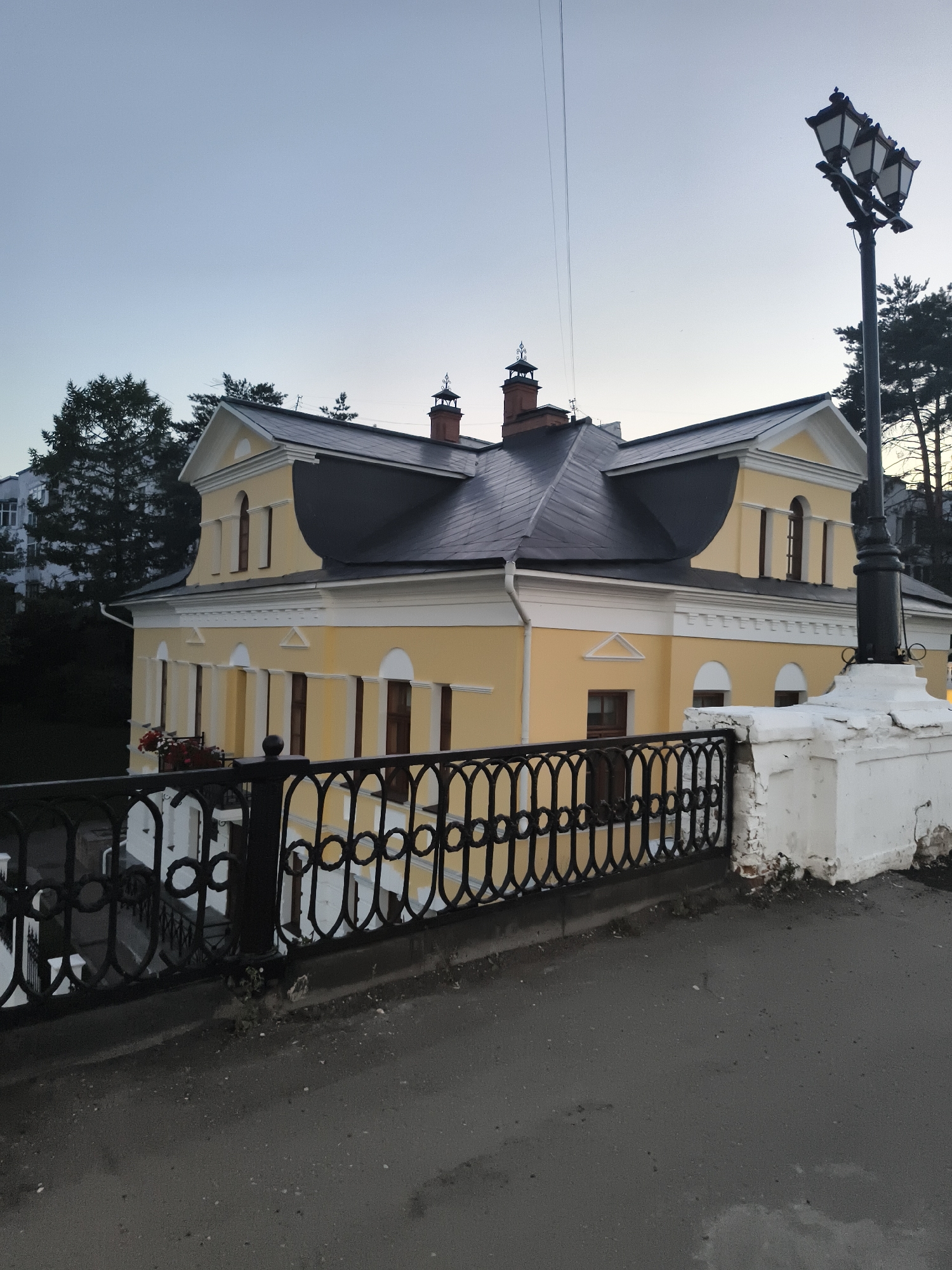This image depicts a yellow building with a dark, multi-pitched roof. The building features several arched windows on the lower level and vertical windows on the upper level. A decorative black metal fence runs along the foreground, separating the building from a paved area. A multi-lamp street light stands on a white pedestal to the right of the building. Trees are visible behind the building and to the left. The sky is a pale blue, suggesting it is either dawn or dusk. The scene appears to be a street or a park area in Yaroslavl, Russia, likely in a historical district given the architectural style of the building and fence. There are no people or animals visible in the image.