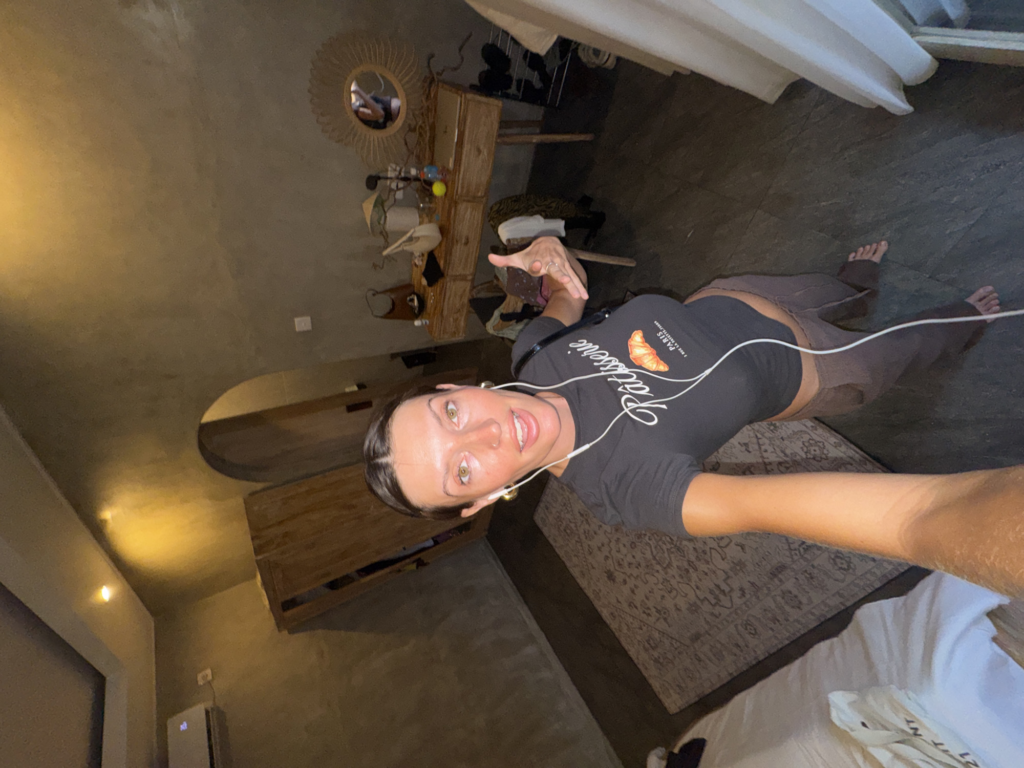 A woman is taking a selfie indoors in North Kuta, Indonesia. She is wearing a black t-shirt with "Patisserie" printed on it, brown leggings, and white earphones. The room contains a bed with white linens, a rug, a dresser with a circular mirror above it, and an air conditioner.