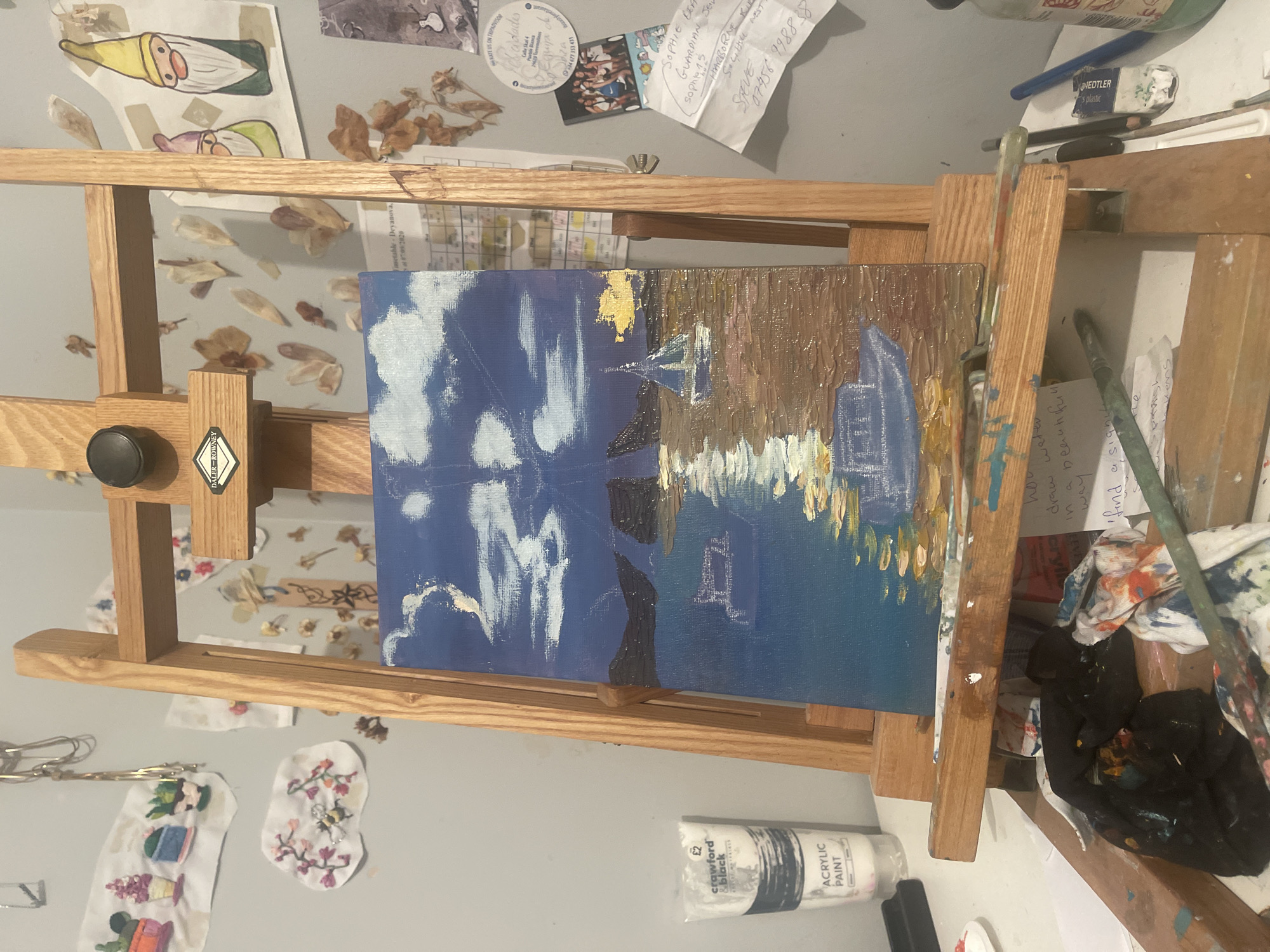 The image depicts an easel holding a canvas painting in progress. The left side of the painting features a blue sky with prominent white clouds, while the right side showcases an impressionistic rendition of buildings and reflections in a body of water, rendered in browns, golds, and blues. Paintbrushes, tubes of paint, and cleaning cloths are scattered around the base of the easel. The background is a light grey wall adorned with various items including drawings of gnomes, dried flowers, magnets, notes and embroidered fabric patches. A tube of "Crawford & Black" acrylic paint is visible. The location in Maidstone, United Kingdom cannot be deduced from the image.