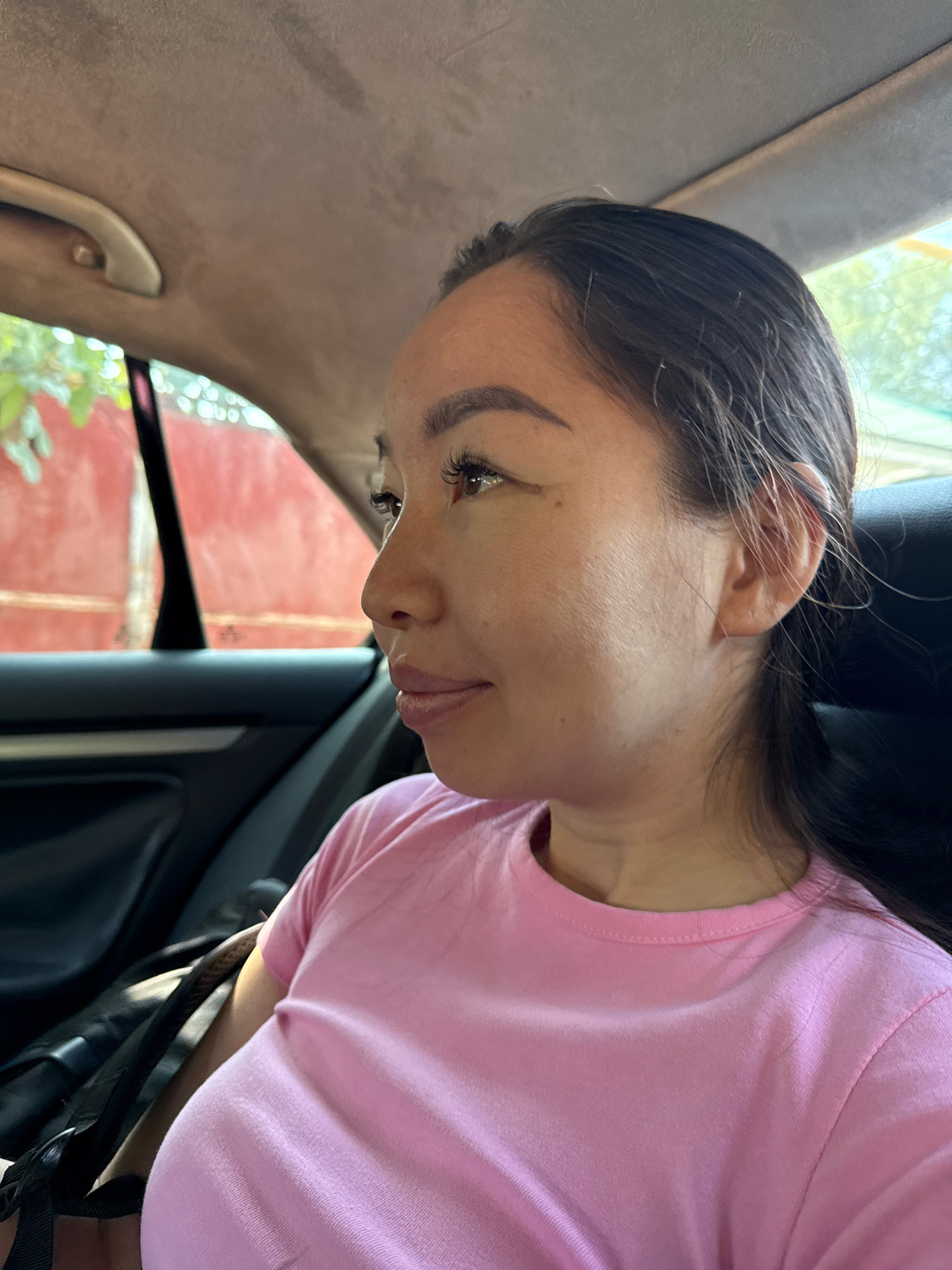 A woman is pictured from the chest up, looking to her right, from inside a car. She is wearing a pink t-shirt. Her hair is dark brown and pulled back. Her makeup includes noticeable eyelashes. The interior of the car is visible, with a grey headliner and black leather seats. The background outside the window shows a blurry red wall and green foliage. The scene appears to be located in the city of Talgar, Kazakhstan.