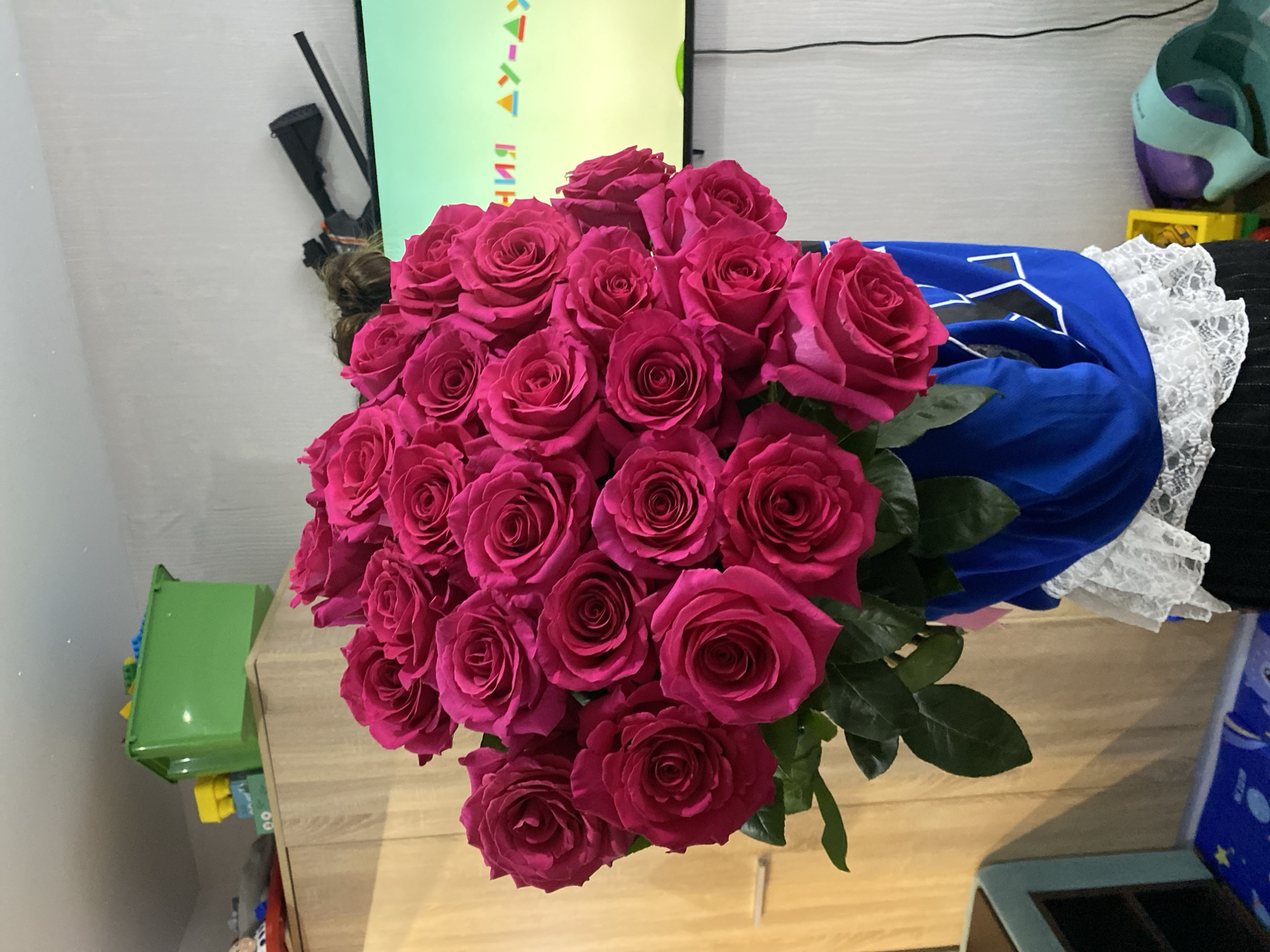 A high-angle shot presents a lush bouquet of deep pink roses. The roses are densely packed, creating a spherical form. Some green leaves are visible. The bouquet is resting on a light-brown wooden surface, which appears to be a tabletop or the top of a piece of furniture.
Behind the roses, there is a glimpse of other items. To the right, a blue cloth with white patterns is partially visible, edged with white lace. Behind the bouquet, and slightly to the left, there's a vertical green object with writing on it. An additional item, a dark-colored device, is situated higher and behind the flowers.
Further to the left, a small, green plastic storage container is present. The location is possibly in Votkinsk, Russia.