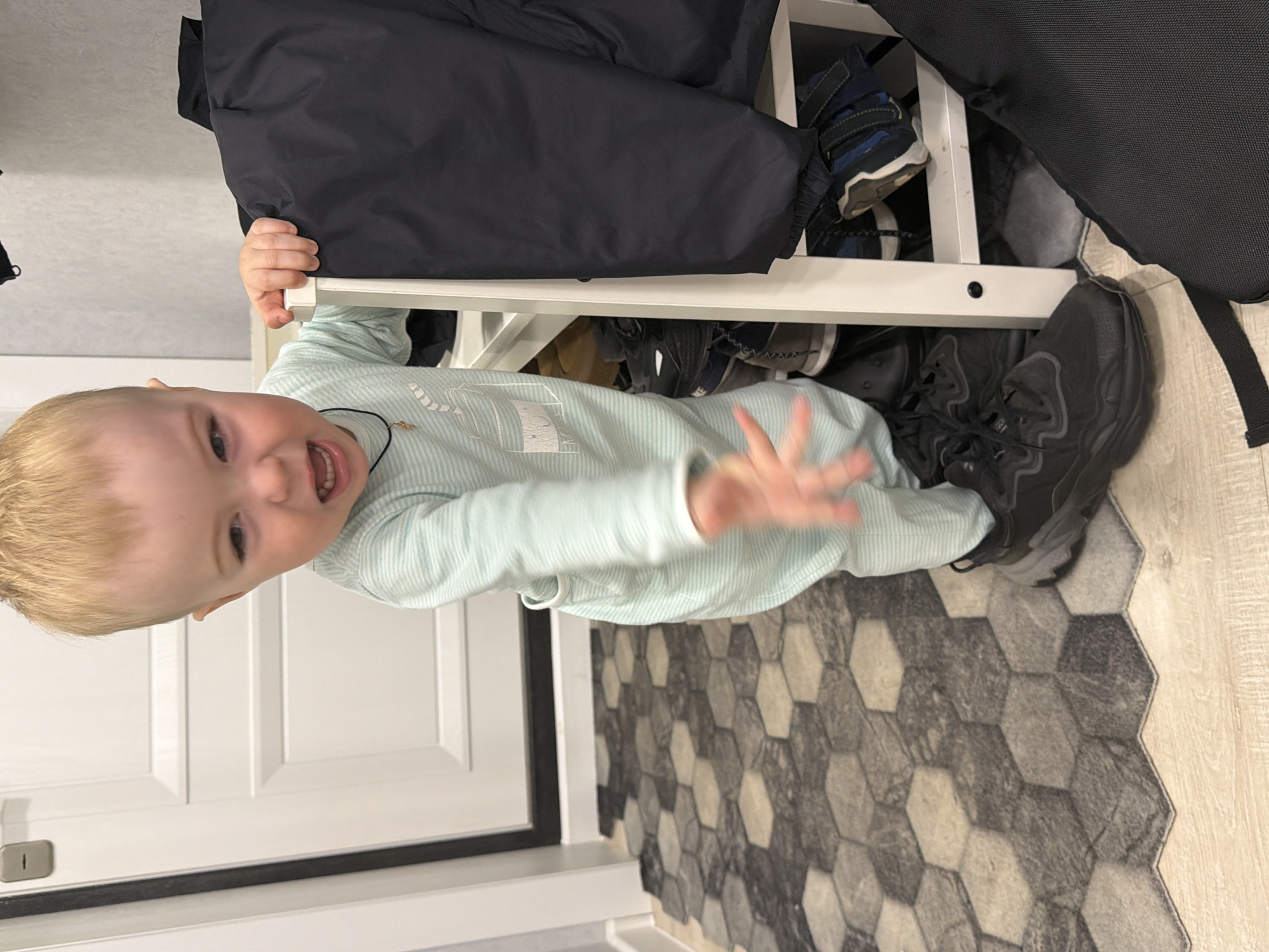 A toddler stands indoors, leaning against a white, two-tiered shoe rack holding multiple pairs of shoes and bags. The child is wearing a light blue striped onesie and is smiling, with his right hand extended. The floor is covered in a patterned rug, and a white door is visible in the background. The location is not discernable from the image.