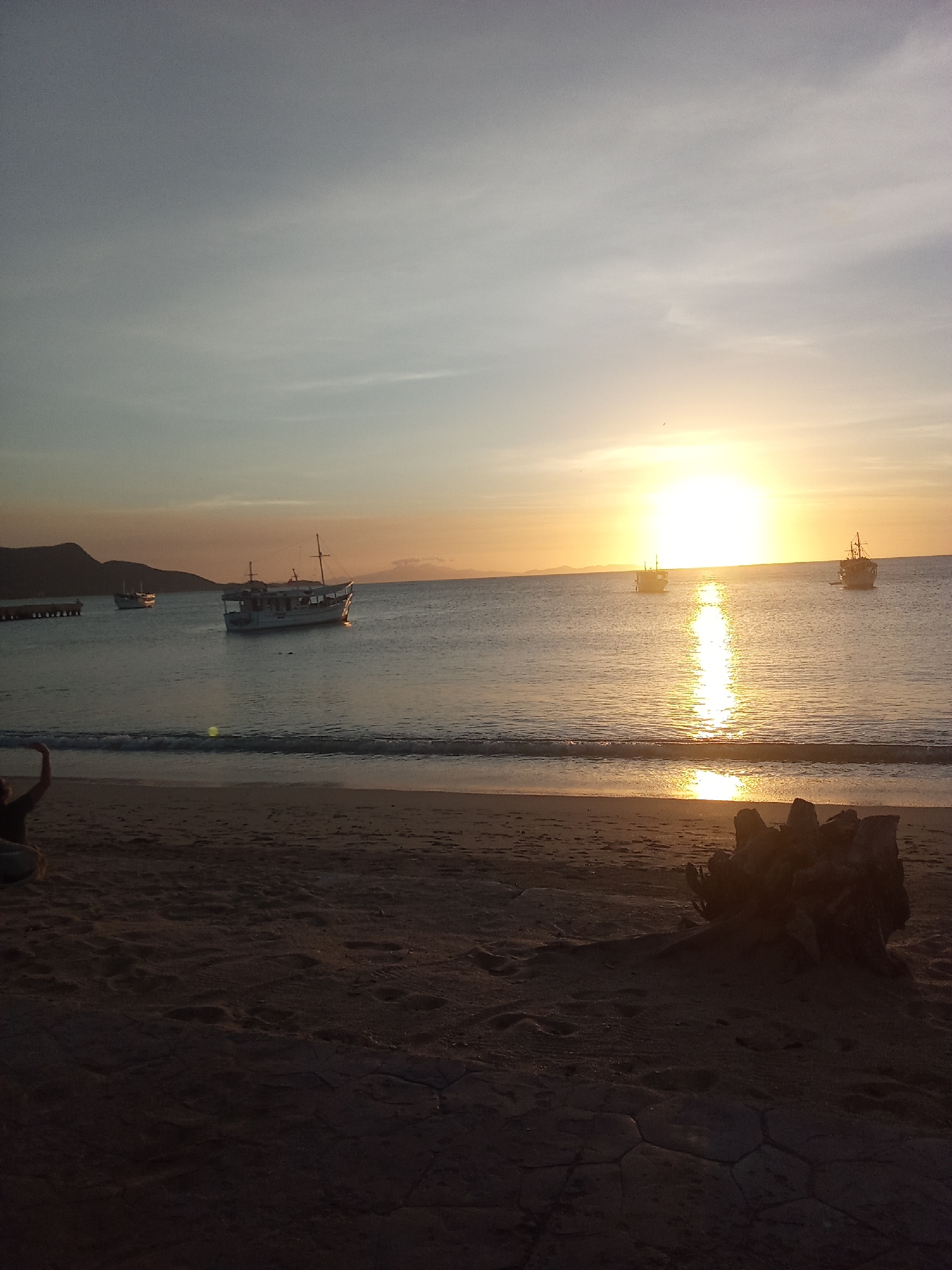 The image depicts a sunset scene at a coastal beach in Juan Griego, Venezuela. The sun, a bright yellow-orange orb, is positioned just above the horizon, slightly right of the frame's center, casting a strong, vertical reflection across the calm, dark blue water towards the sandy foreground. The sky transitions from light blue-grey at the top to warm orange and yellow tones near the horizon, with subtle cloud formations. In the mid-ground, four boats are visible on the water. One prominent white vessel with a cabin and mast is positioned left of the sun's reflection. A smaller, darker boat is further left, near a dark, multi-pillared pier extending from a distant landmass on the far left. Two additional boats, appearing as dark silhouettes, are located further out on the right side of the frame, one directly within the sun's reflection. Distant hills or mountains are silhouetted against the horizon on both sides of the bay. The immediate foreground consists of dark sand, showing footprints, and transitions to a darker, patterned ground surface, possibly cracked earth or paving, at the very bottom. On the far left of the foreground, the partial silhouette of a person is discernible, with their left arm raised and bent at the elbow. On the right side of the foreground beach, a dark, irregularly shaped object resembling a tree stump or roots is present. Small waves are breaking softly on the shoreline.
