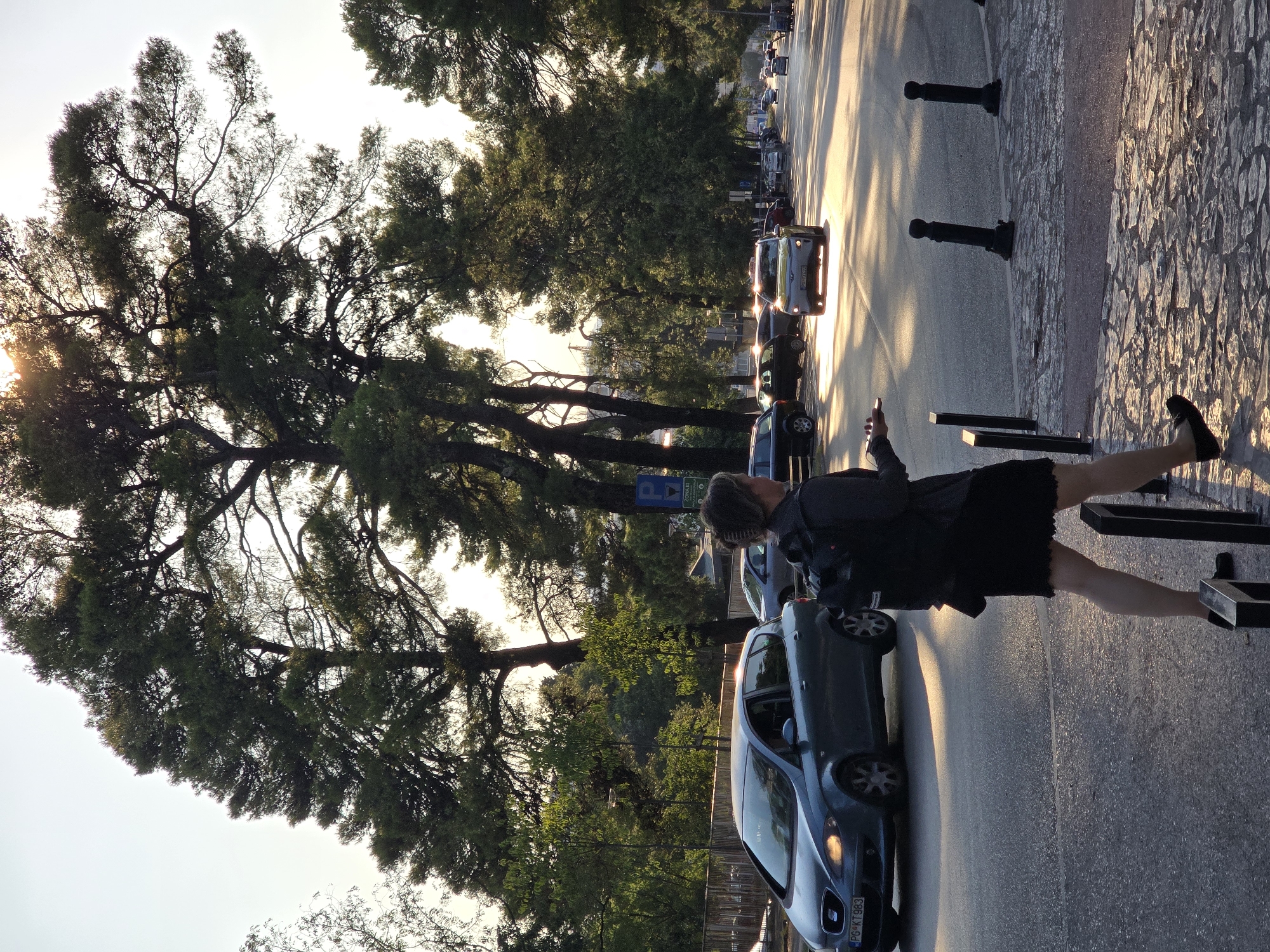 A medium shot shows a woman walking on a pedestrian area next to a street in Podgorica, Montenegro. The woman is in the foreground, facing away from the camera, and is dressed in a dark skirt and a grey jacket. She has a backpack and is holding a mobile phone. The street is lined with tall trees, and the setting sun casts long shadows. Several cars are driving on the street, some of which are illuminated by the sunlight. A parking sign is visible on the right side of the image. The ground in the foreground is paved with stones.