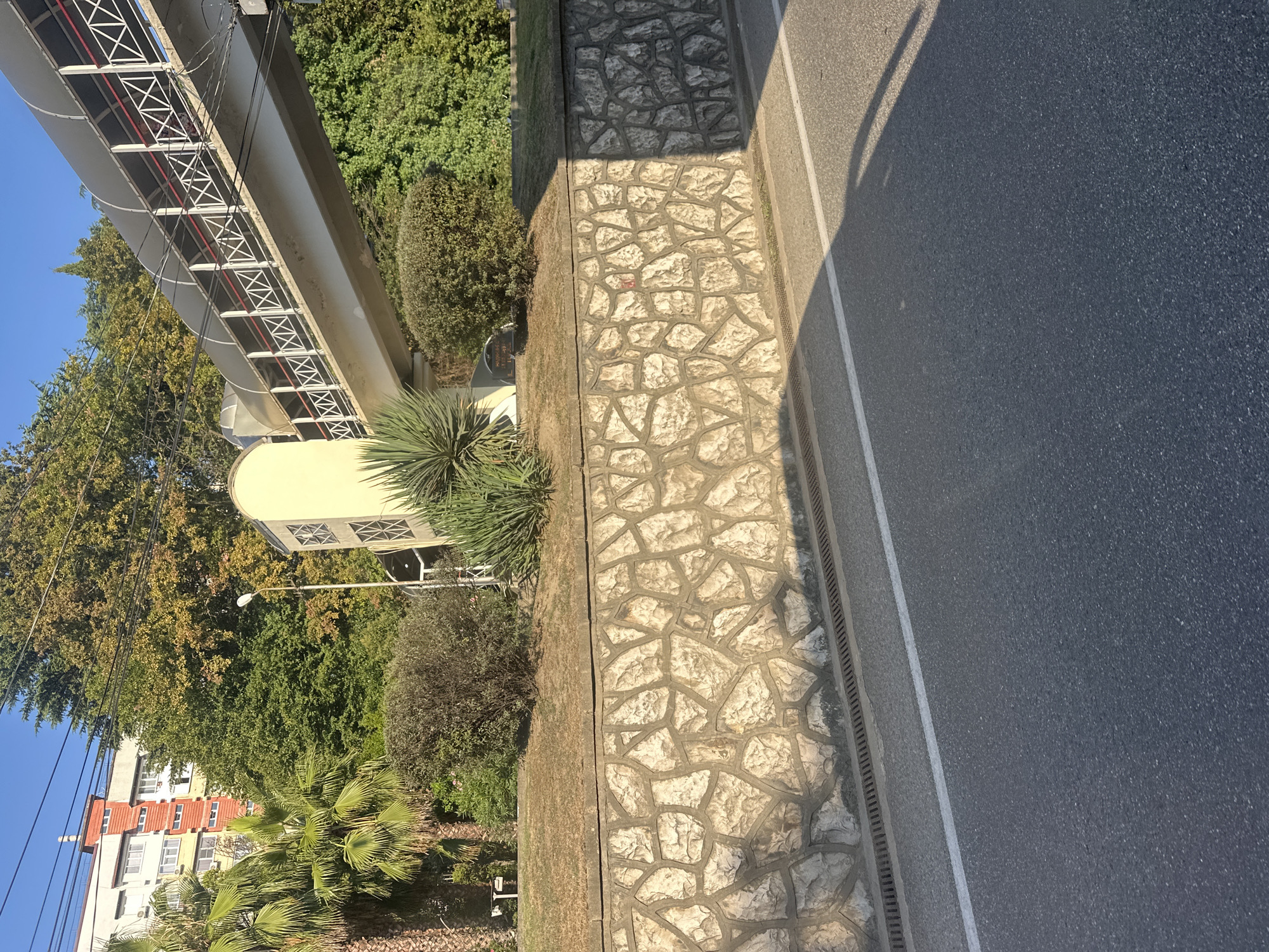 The image depicts a pedestrian overpass in Sochi, Russia, likely located near the coast given the presence of palm trees and lush greenery. The overpass, a long, elevated structure with a covered walkway, spans across a roadway. It features a white railing with red accents and a translucent canopy. At the base of the overpass, there is a light-colored, curved structure. Adjacent to the overpass, a multi-story building with a red and white facade is visible, along with dense trees and shrubs. A stone retaining wall, constructed from irregularly shaped stones, supports a grassy embankment. The roadway in the foreground is paved and marked with a white line. The scene is bathed in sunlight, casting shadows from the overpass onto the road.