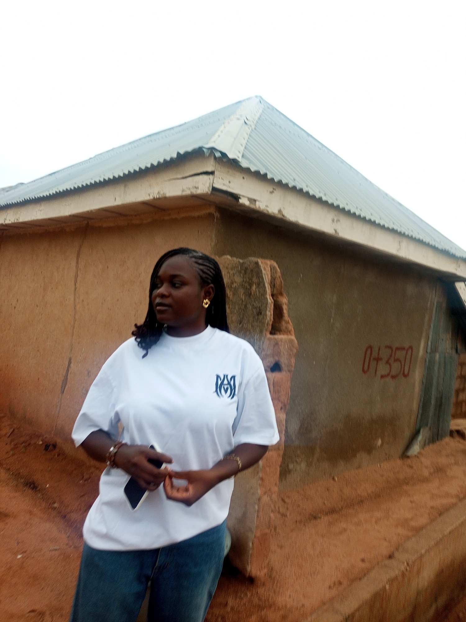 A young, dark-skinned woman stands outdoors in what appears to be Tempure, Nigeria. She has braided hair with gold earrings and wears a white t-shirt with a dark emblem on the left breast and jeans. She holds a dark-colored smartphone in her right hand and gestures with her left.
Behind her, a small, weathered building with a corrugated metal roof is visible. The building has light brown walls with the faded inscription "0+350" in red lettering. The ground around the building is reddish-brown soil. The sky is overcast.