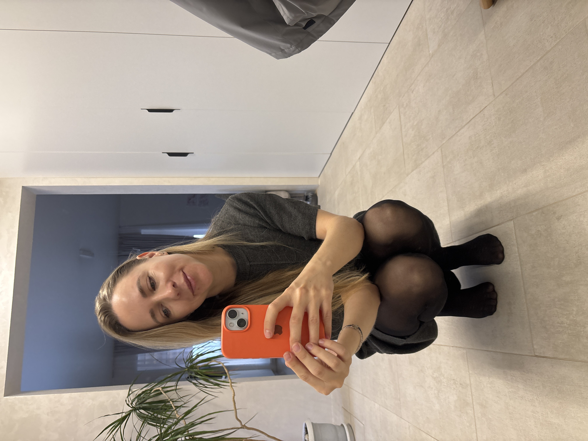 A woman with blonde hair is crouched on a light-colored tiled floor, holding an orange iPhone in front of her. She is wearing a gray dress and black tights. Behind her is a white wall with a doorway leading to another room and some potted greenery. The room has white cabinets and a light-colored tiled wall. The location is Chaikovsky, Russia.