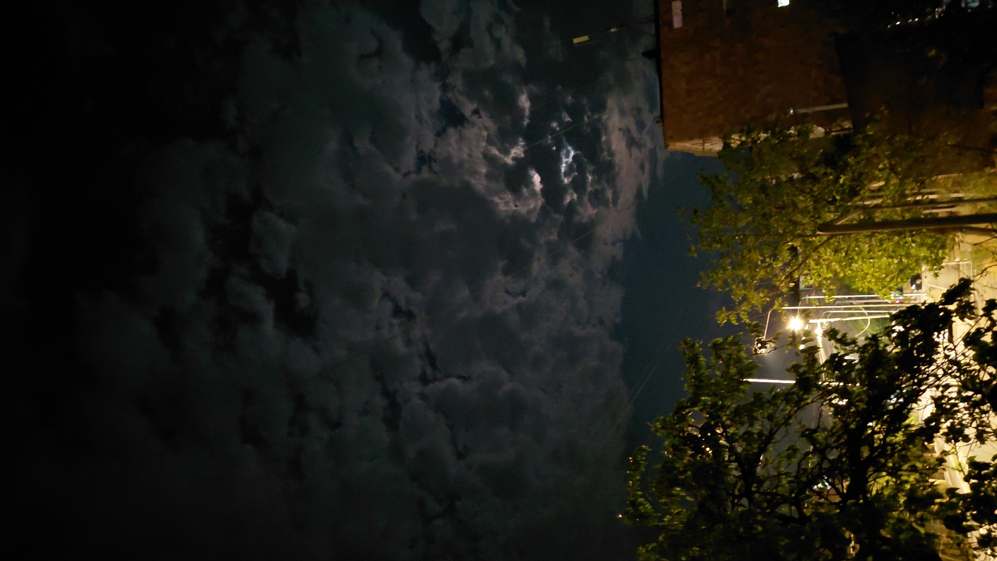 This is a nighttime, low-light, vertical shot of a partially cloudy sky over Abovyan, Armenia. The sky is dark with scattered clouds, some of which are illuminated by the moon, creating a textured, mottled effect. In the lower portion of the image, a building with a brick facade is visible on the right, with a dark roofline. To the left of the building, trees with green foliage are silhouetted against the sky and streetlights. A street with several streetlights is visible, with some subtle indications of traffic or movement. The overall impression is of a quiet, urban night scene.